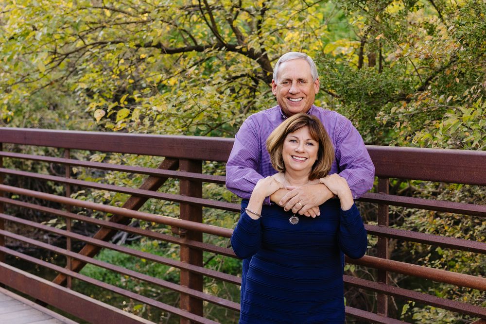 A couple embraces while standing on a wooden bridge surrounded by autumn foliage in the background.