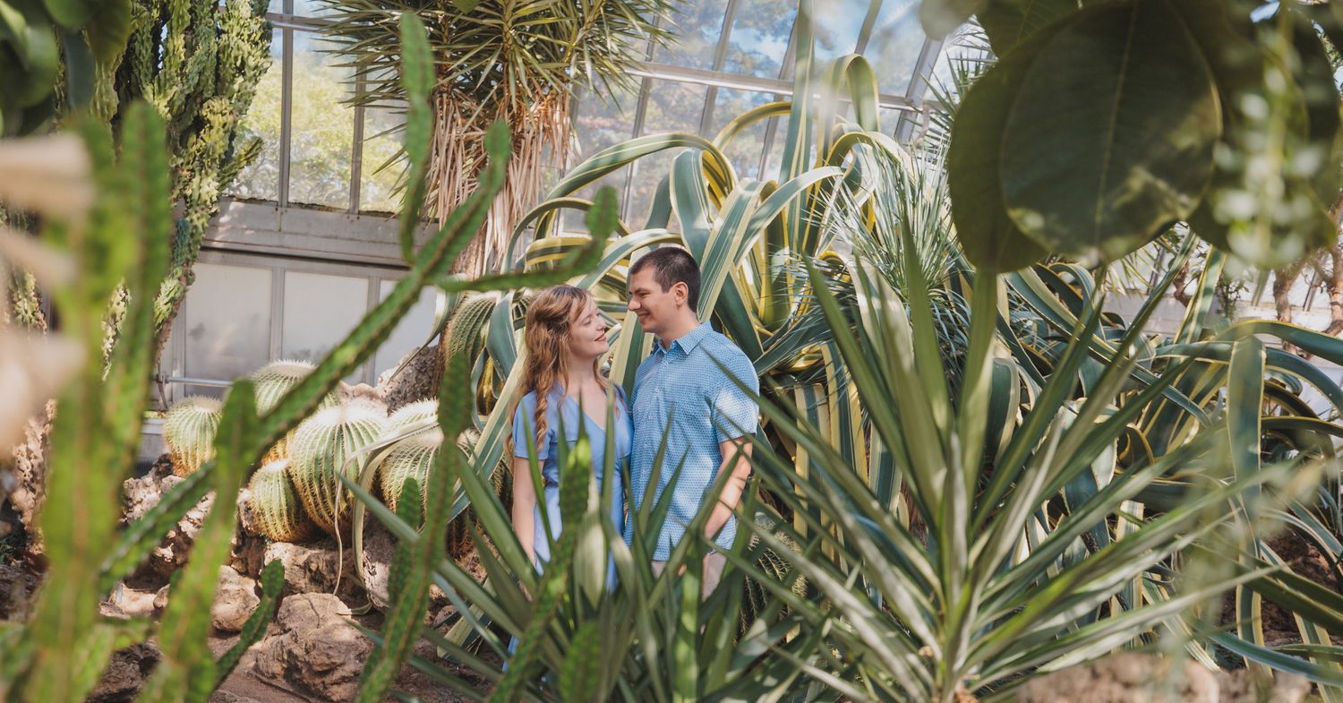 Two people walk through an indoor greenhouse filled with tall cacti and tropical plants on display.