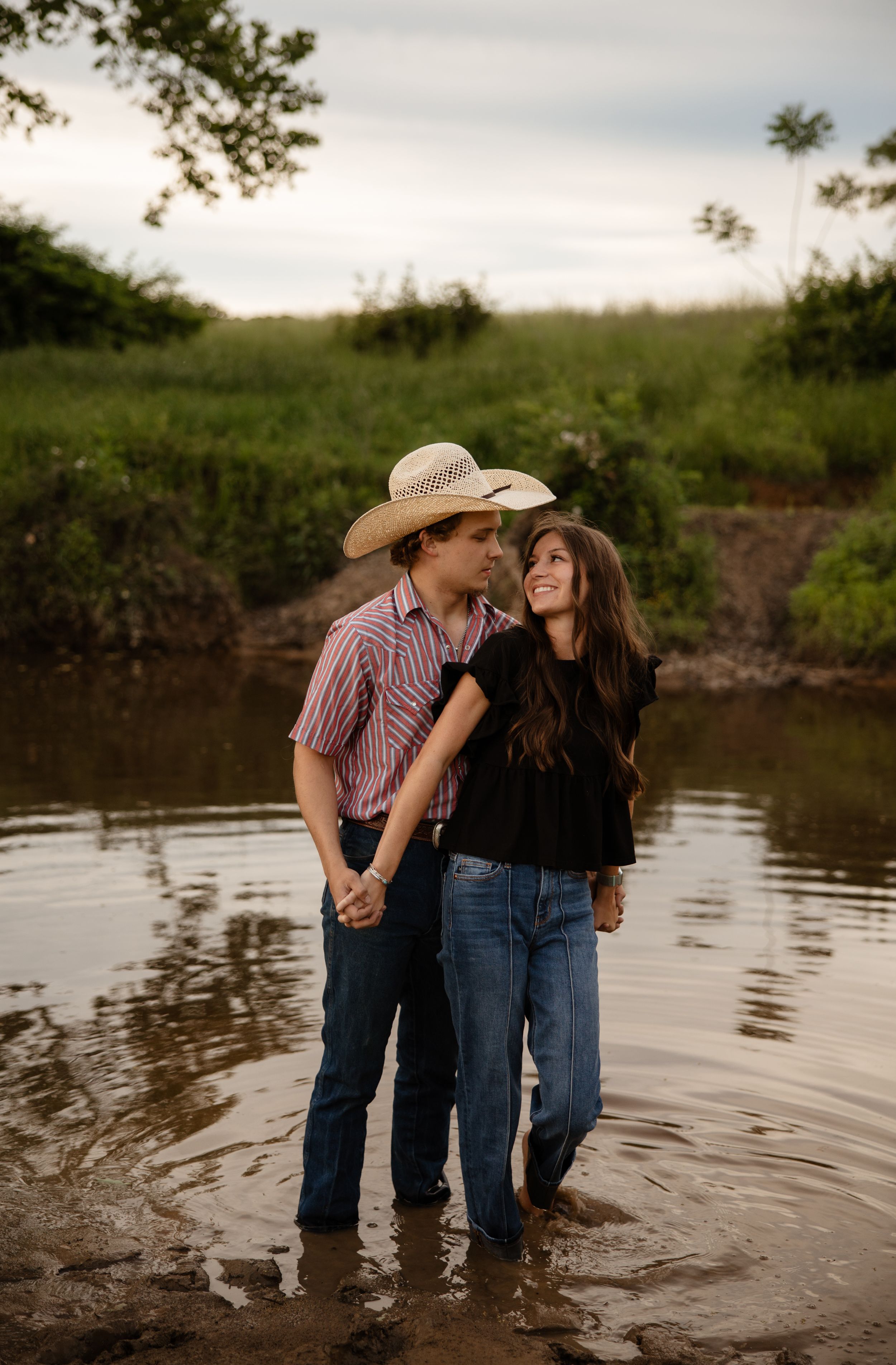 A romantic couple stands ankle-deep in a pond at sunset, sharing an intimate moment in a rustic country setting.