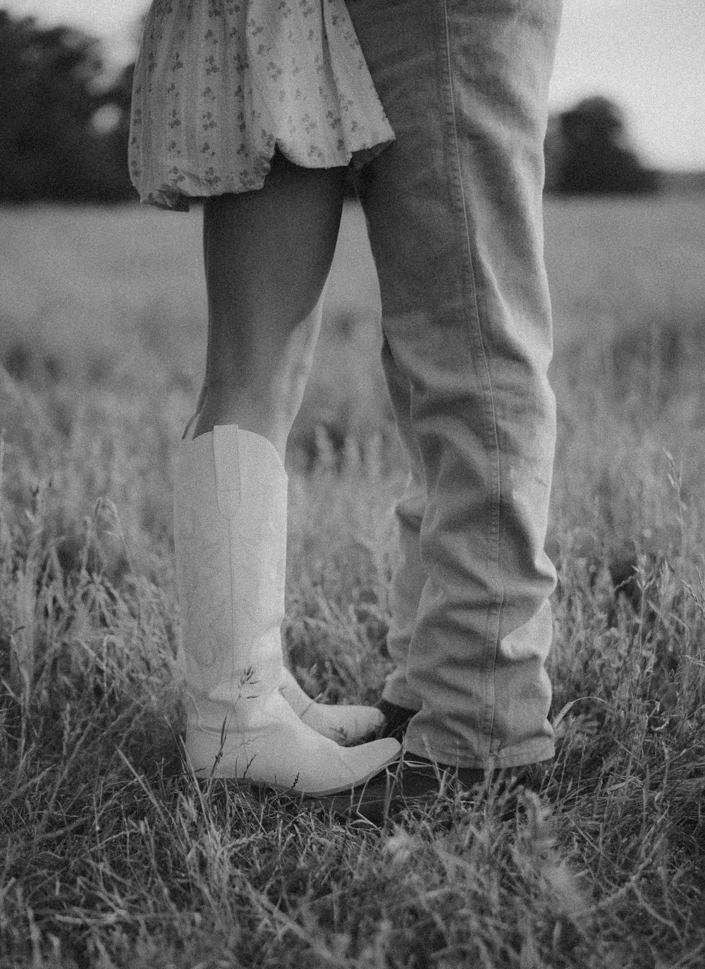 A close-up black and white photo shows two pairs of cowboy boots standing together in a grassy field.