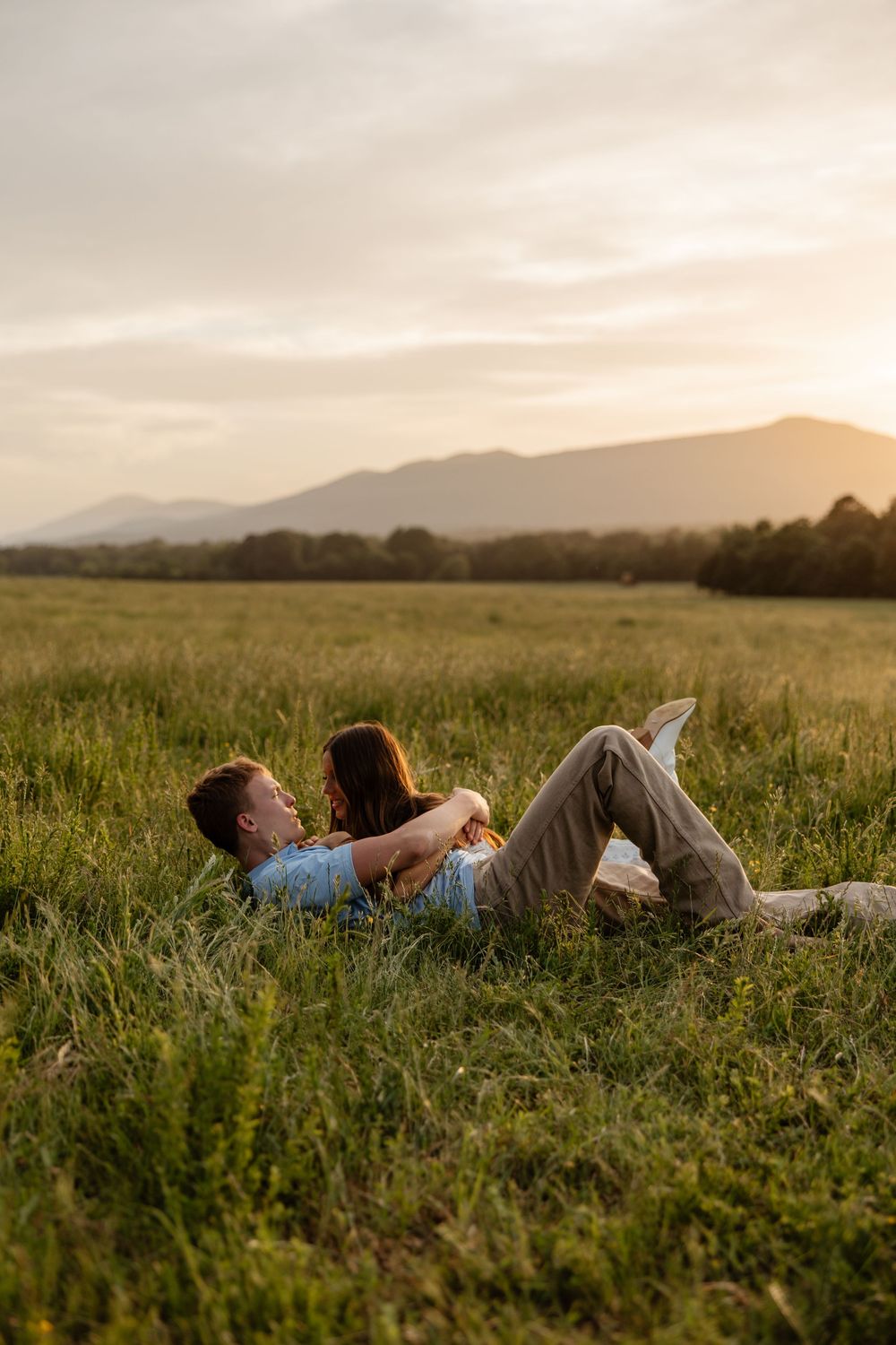A couple lying together in a grassy meadow at sunset with mountains in the background.