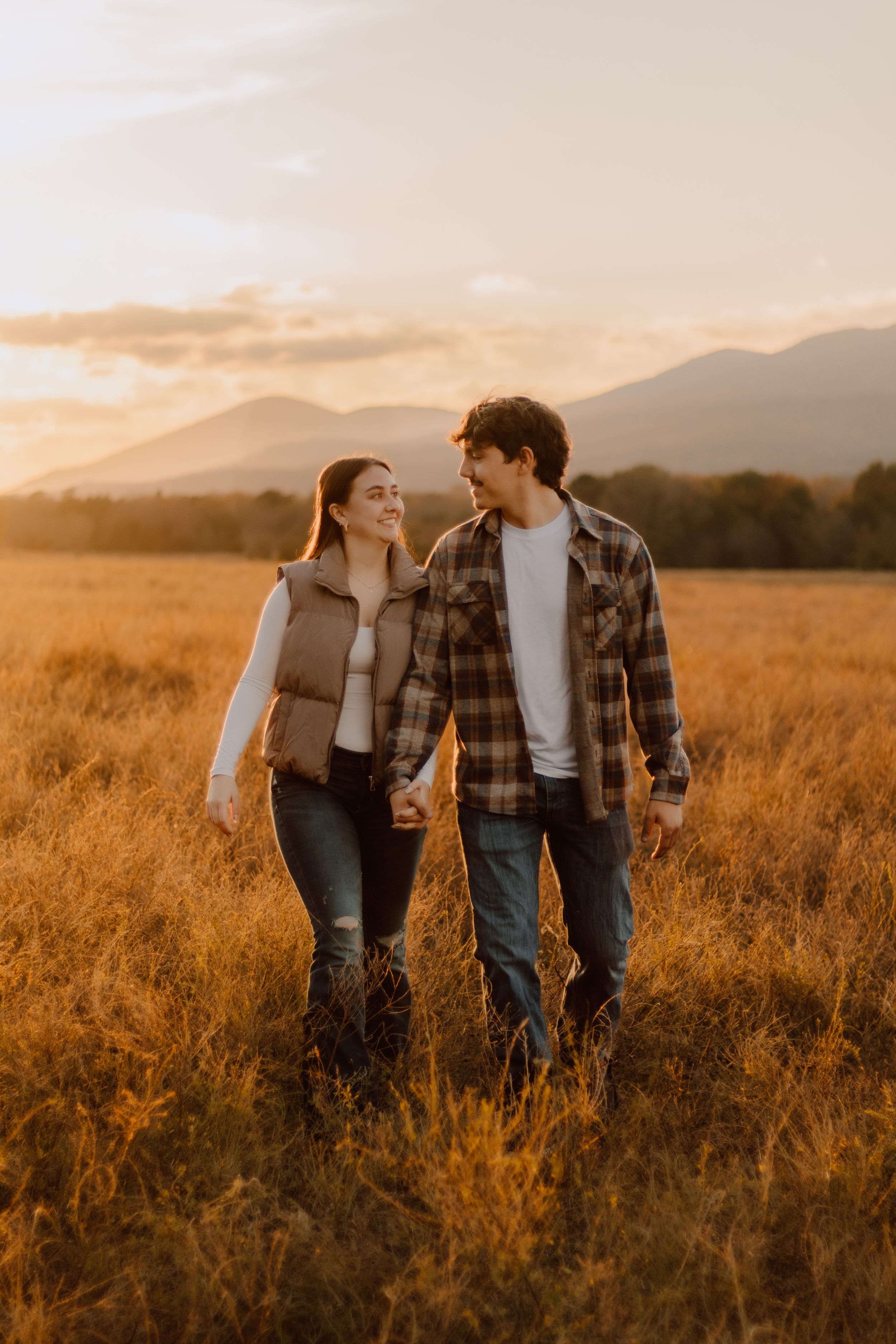 Couple in plaid shirts walking together in a golden wheat field at sunset with mountains in the background.