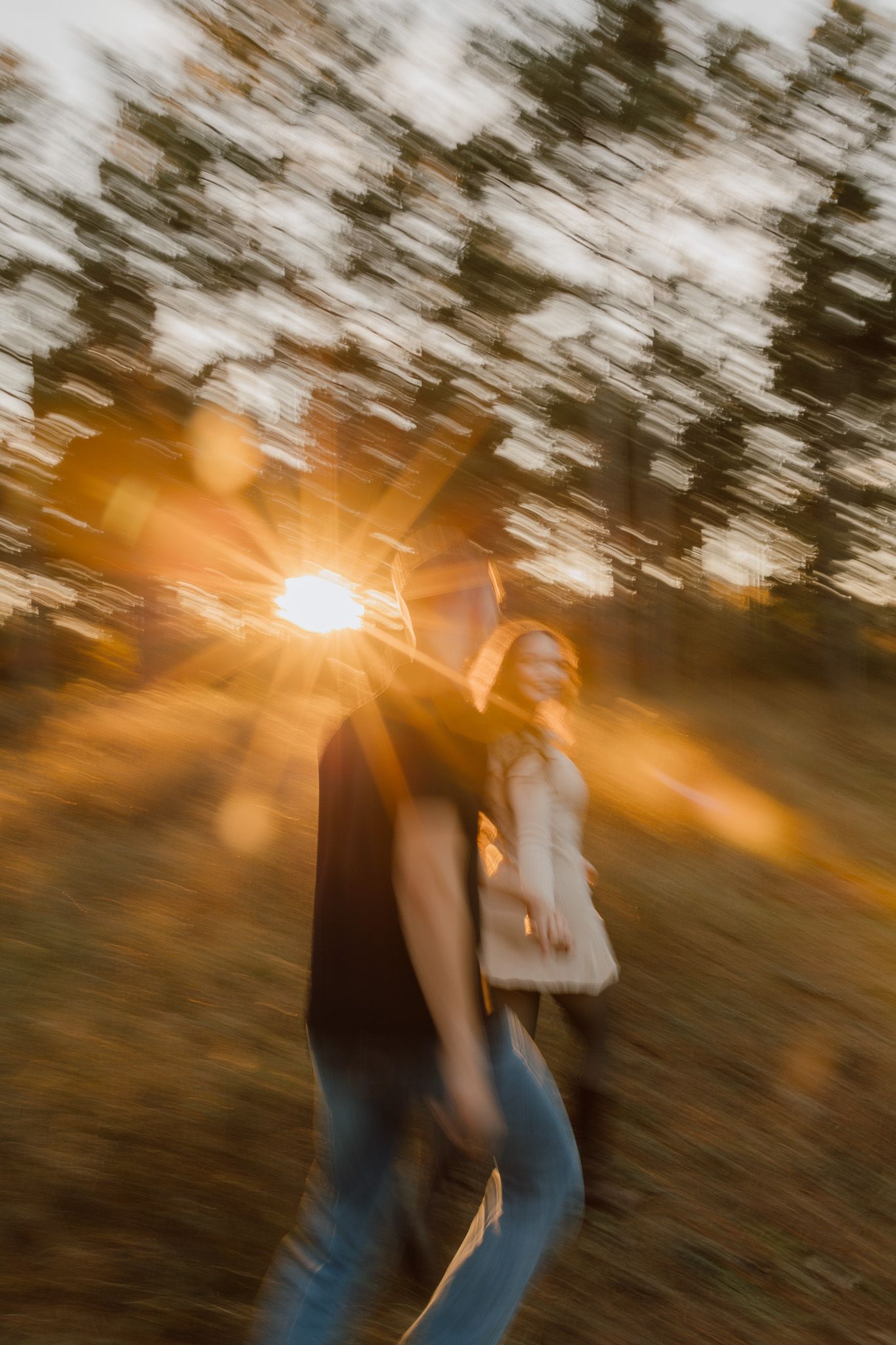 A couple embraces in motion during golden hour sunset with sun flares and trees creating a dreamy, romantic atmosphere.