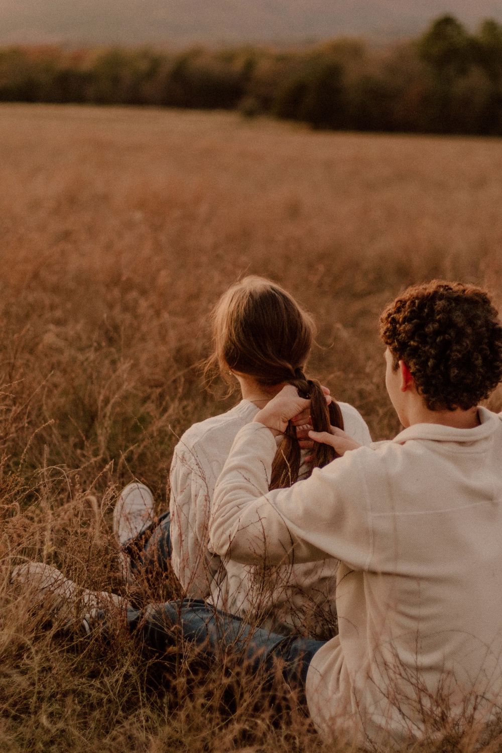 A couple sits together in a golden field during sunset while braiding hair.