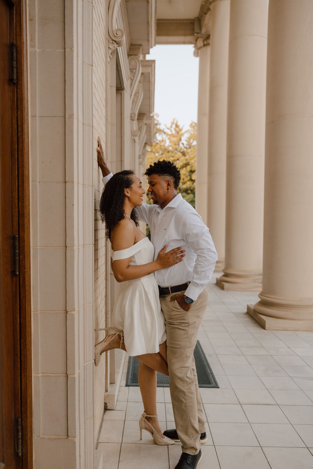 A romantic couple shares an intimate moment in an elegant colonnade hallway with white pillars and warm lighting.