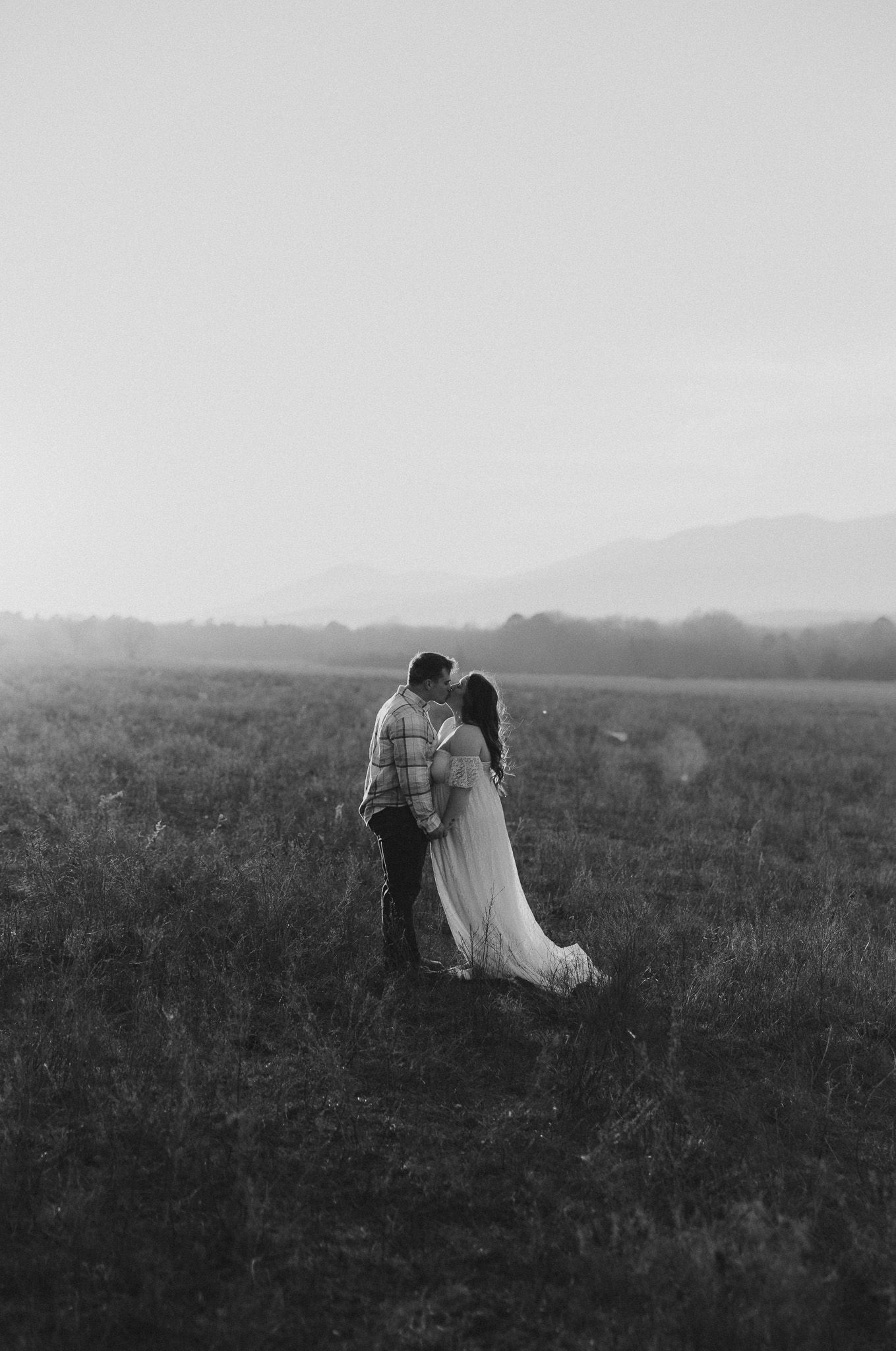 A couple in wedding attire walks together through a misty field at dusk in black and white photography.