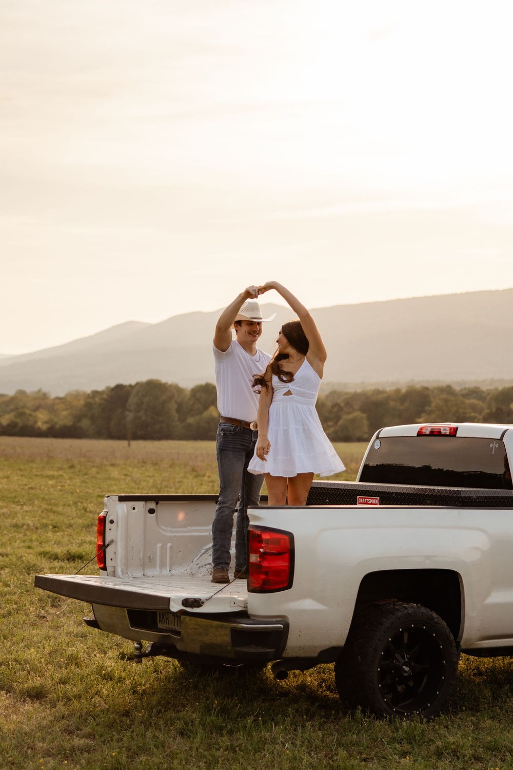 A couple dances together on the back of a white pickup truck in a scenic field during golden hour.