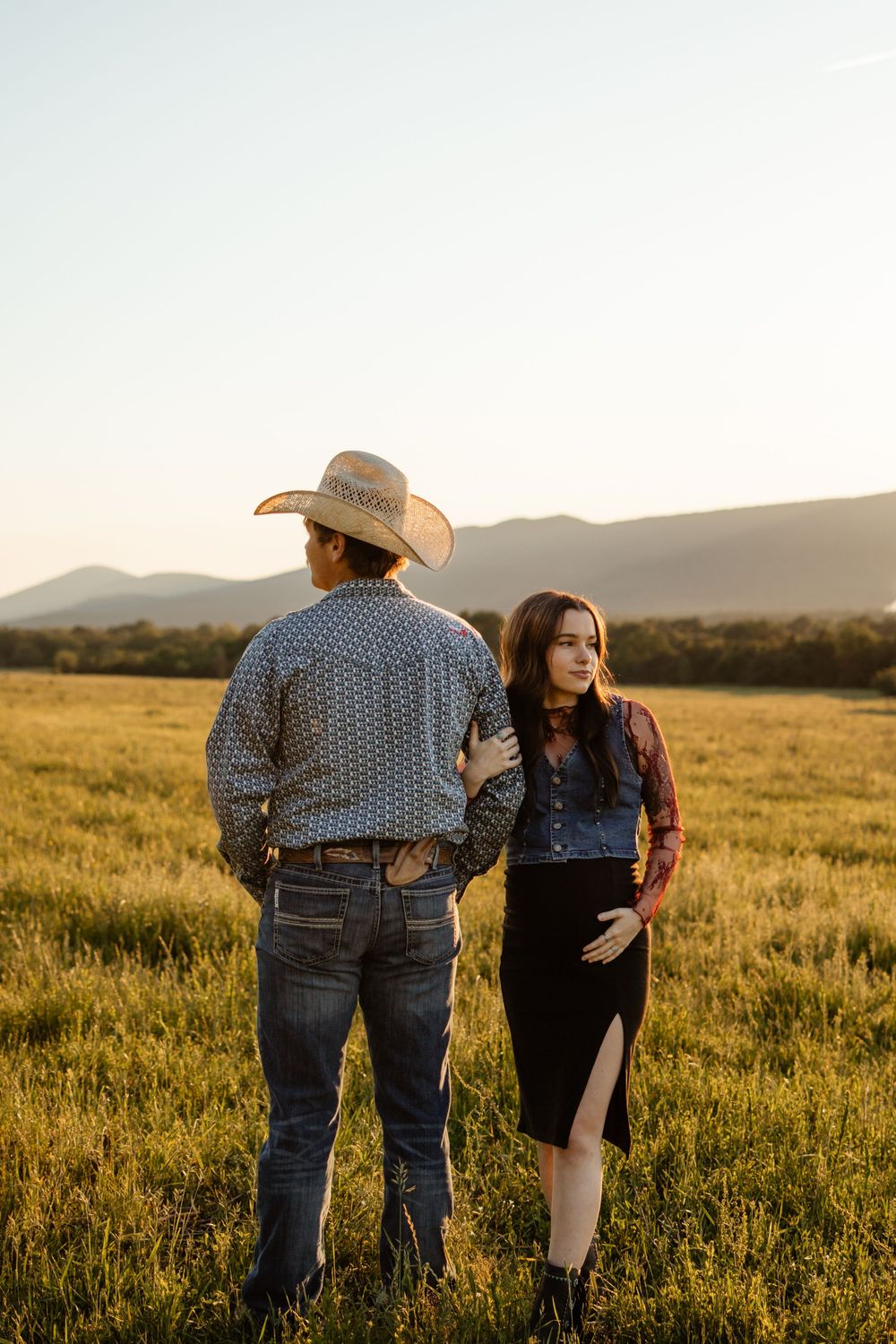 A couple walking through a golden grass field at sunset wearing western attire and cowboy hats.