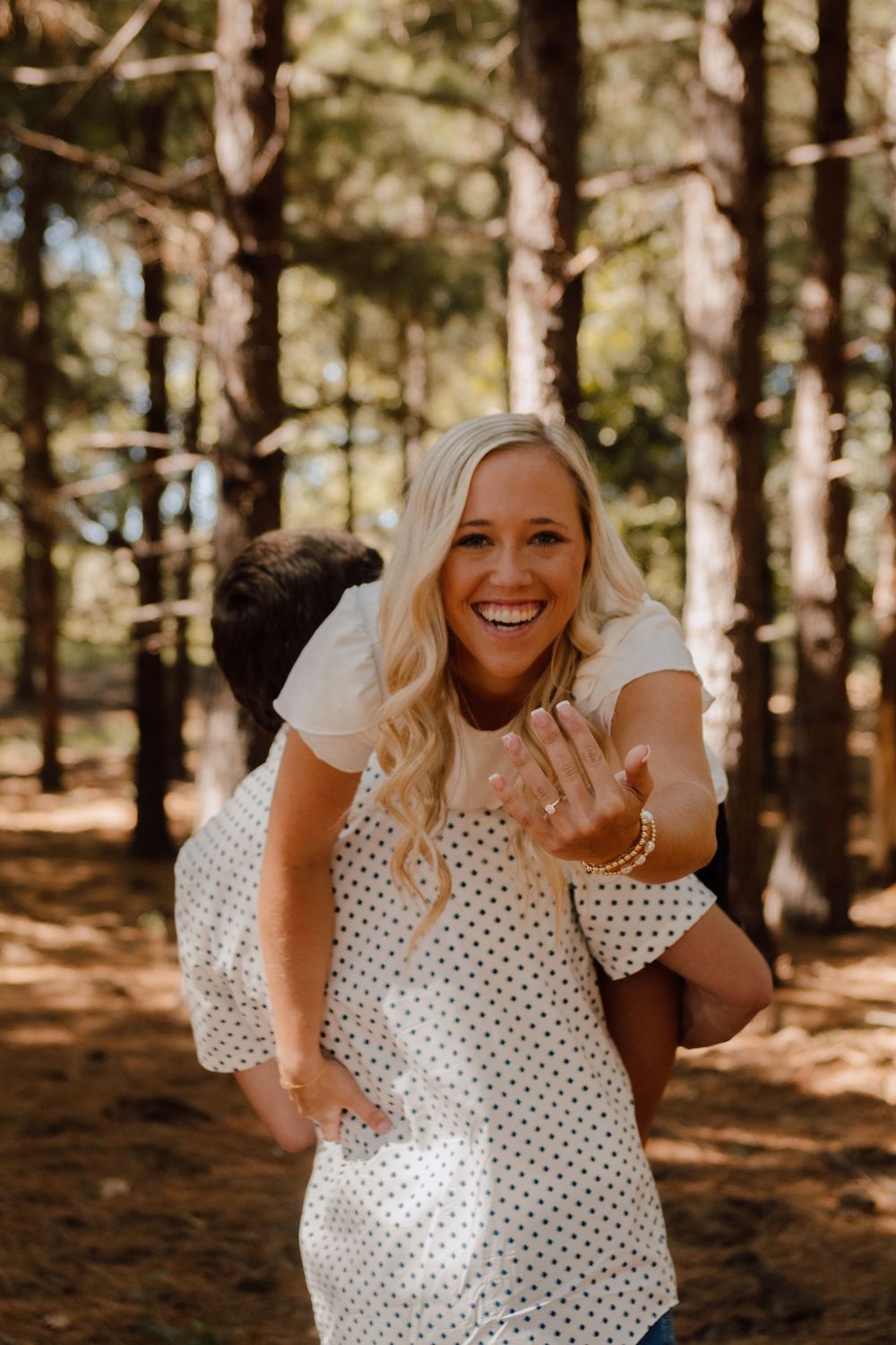 A person in a polka dot dress shows off an engagement ring in a sunny forest setting.