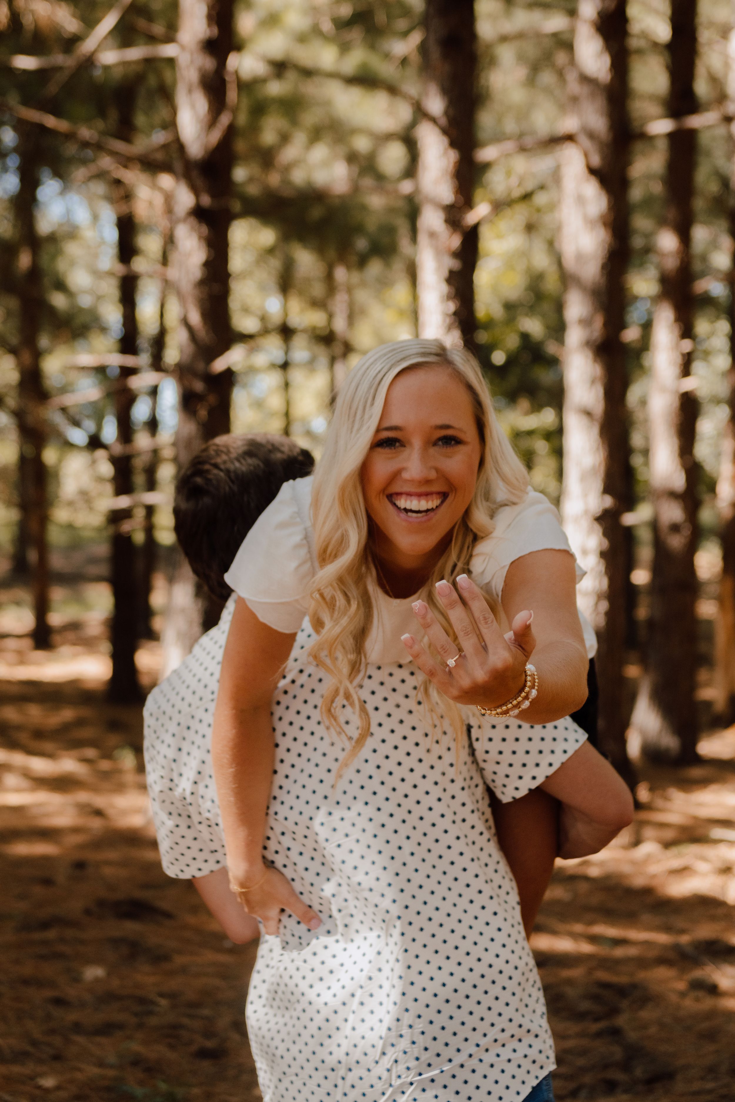 A person in a polka dot dress shows off an engagement ring in a sunny forest setting.