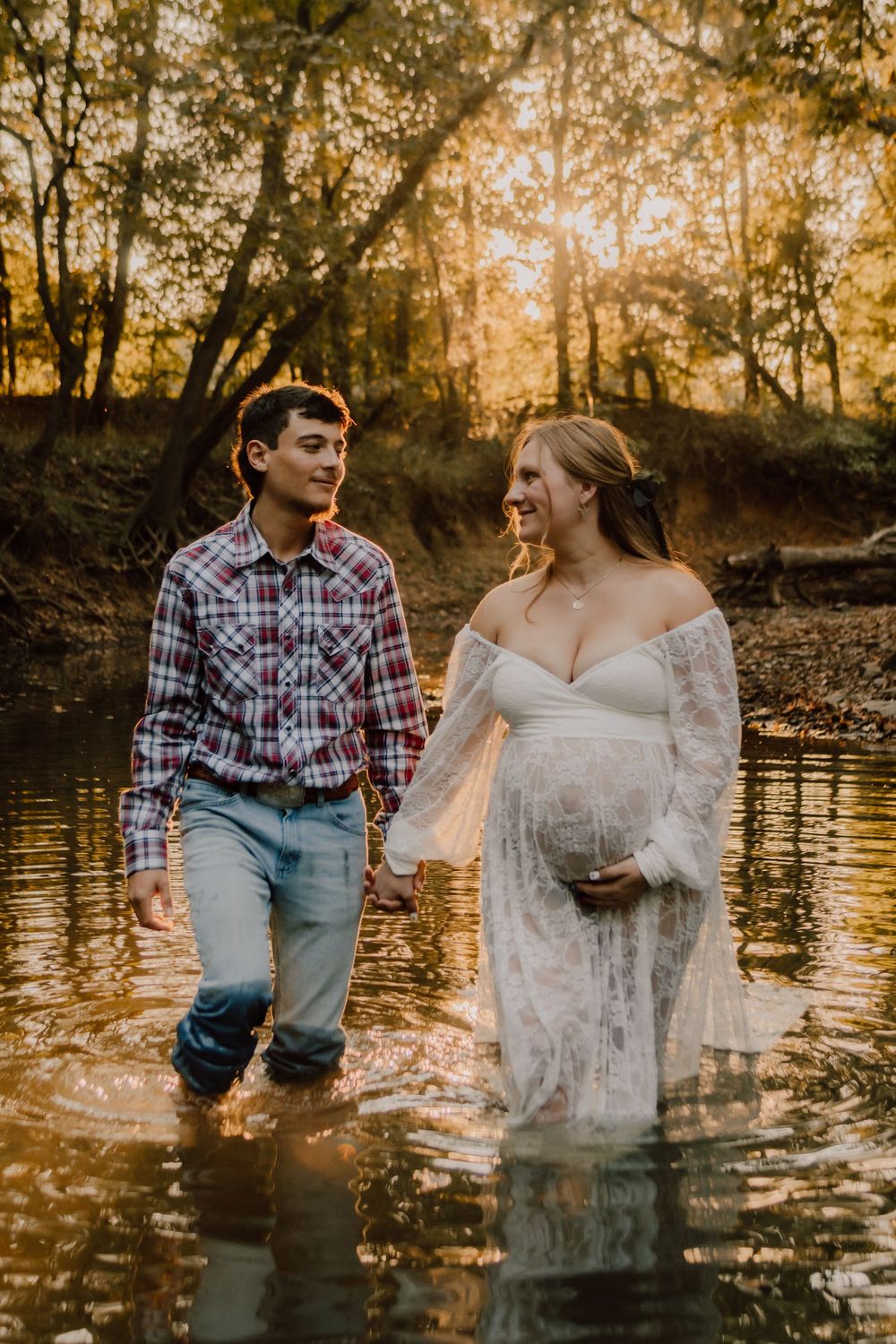 A couple stands together in shallow creek water during golden hour sunset in a wooded forest setting.