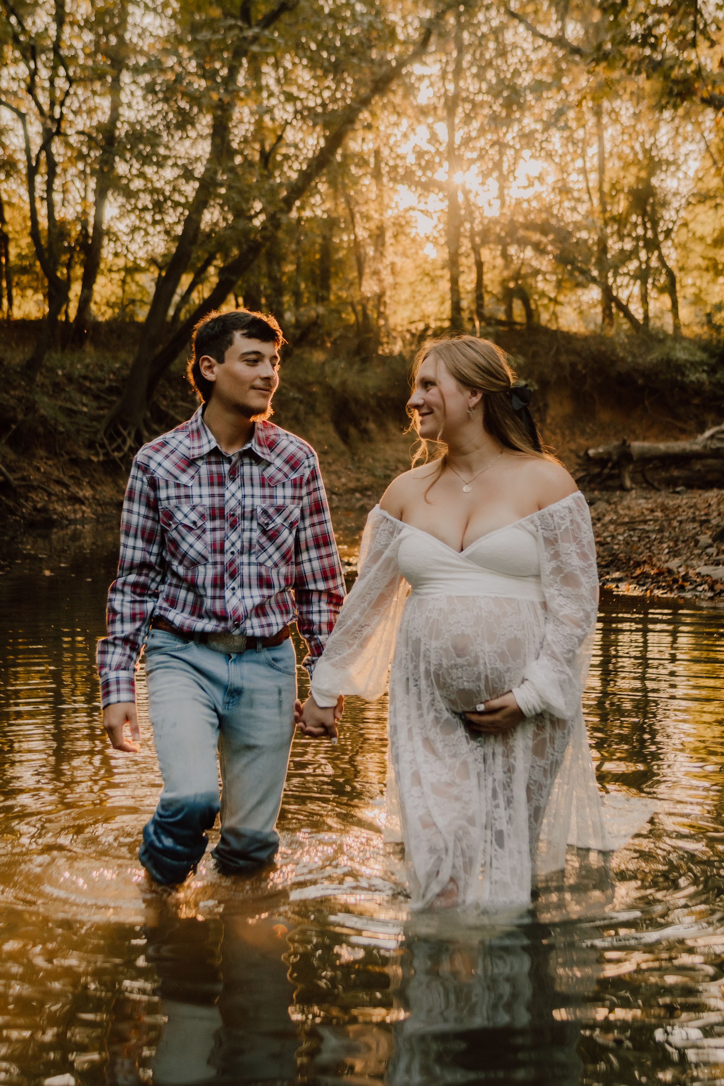 A couple stands together in shallow creek water during golden hour sunset in a wooded forest setting.