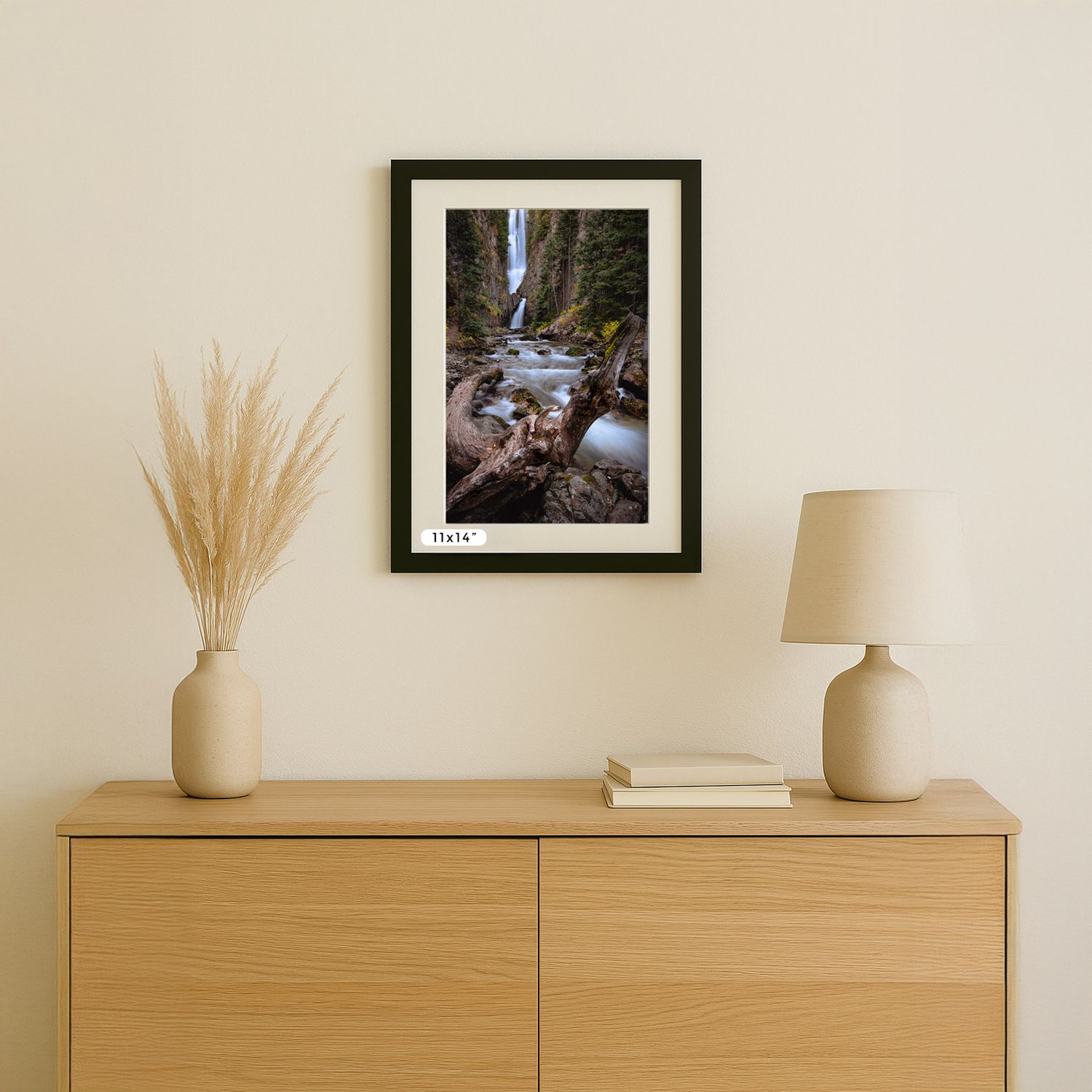 A framed waterfall photograph displayed on beige wall above wooden cabinet with decorative pampas grass and table lamp.