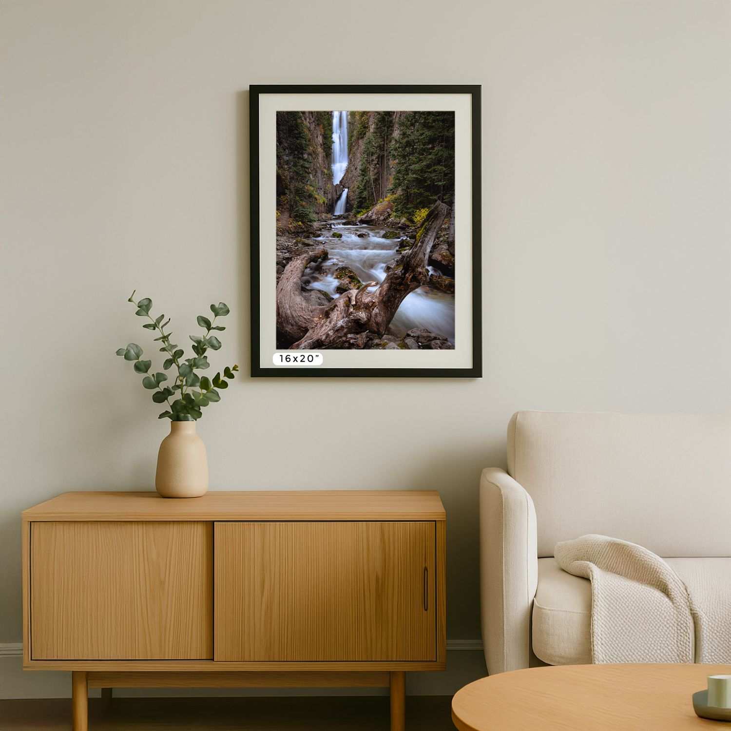 Framed waterfall photograph displayed above wooden credenza with potted plant and partial view of beige armchair.