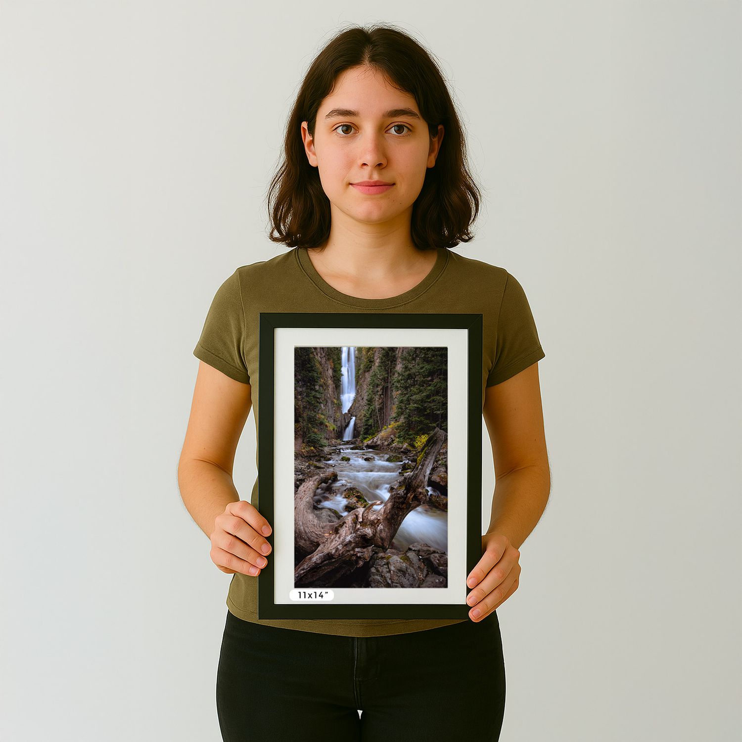 A person in an olive t-shirt holds a framed nature photograph showing a waterfall cascading through rocks.
