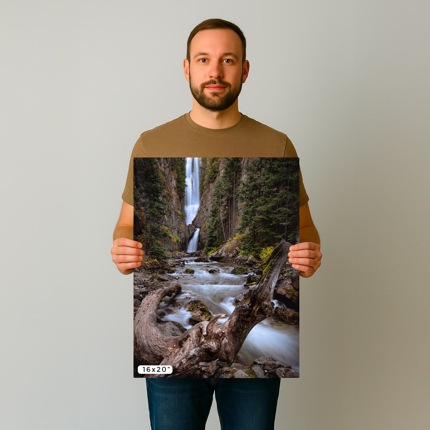 Someone holding a framed nature photograph showing a cascading waterfall and rocky stream through a forested canyon.