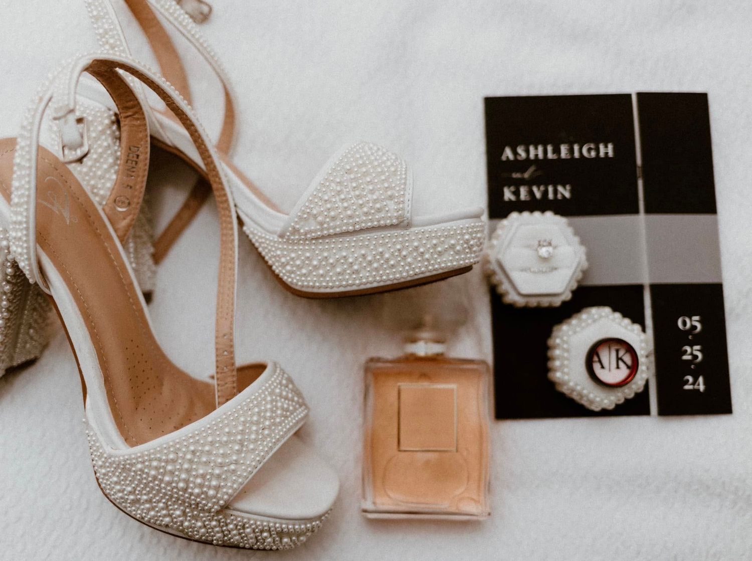 Elegant bridal accessories including white lace heels, perfume bottle, and black invitation card arranged on white fabric.