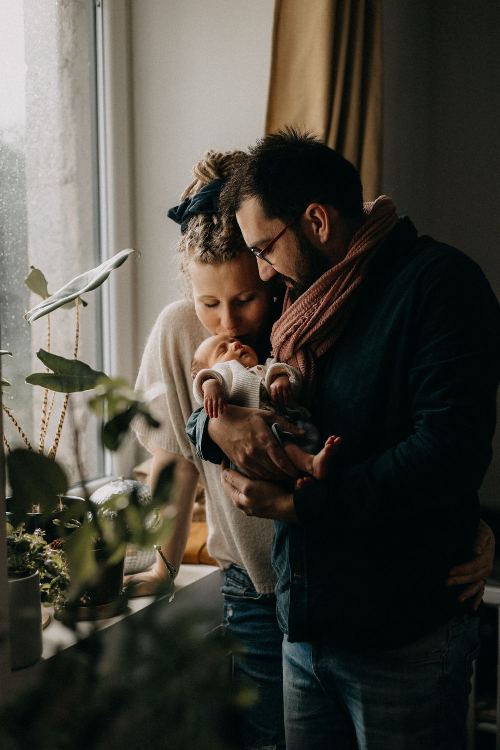 A cozy indoor scene by a window with houseplants as a couple shares an intimate moment with their newborn.