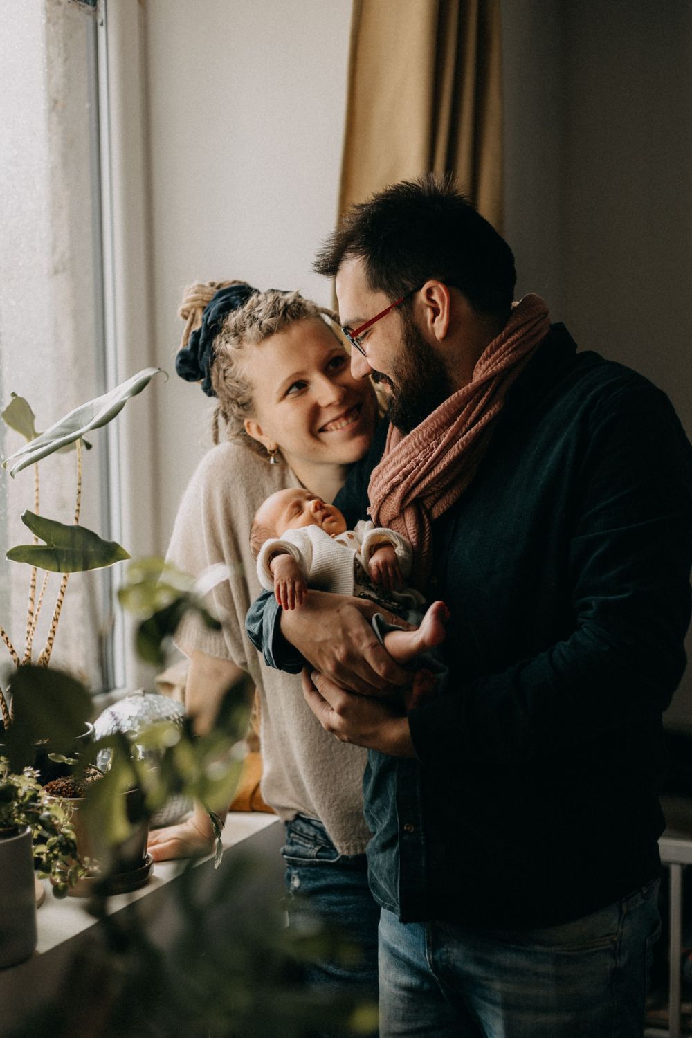 A tender family moment captured by a window with natural light streaming in as parents bond with their baby.