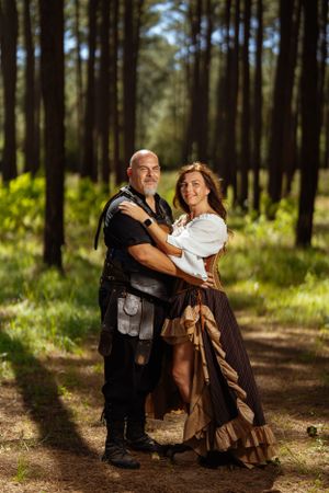 A couple in medieval fantasy costumes poses together on a forest path surrounded by tall pine trees.