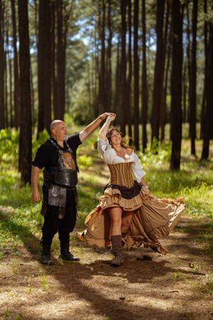A couple in medieval renaissance costumes dancing together in a sunlit forest setting with tall pine trees.