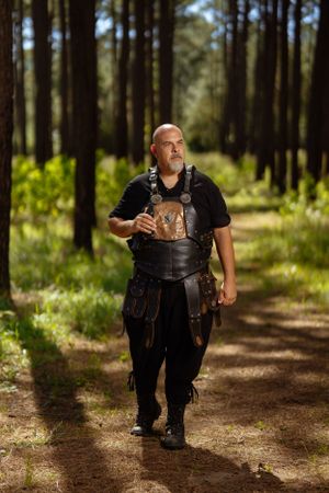 A warrior in black leather armor walks through a sunlit forest path carrying medieval-style equipment.