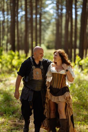 A costumed couple in medieval and renaissance attire share a moment while walking through a pine forest.