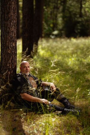 A costumed warrior in black leather armor rests against a tree in a sunlit forest clearing.