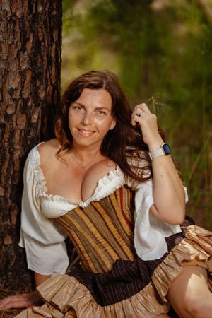 A woman in Renaissance-style dress with a brown corset poses by a tree in a forest setting.