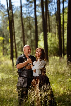 A costumed couple in medieval attire shares a romantic moment while standing in a wooded forest clearing.