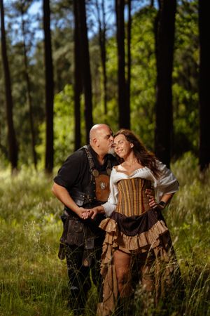 A loving couple in medieval Renaissance-inspired costumes share a tender moment in a sunlit forest clearing.