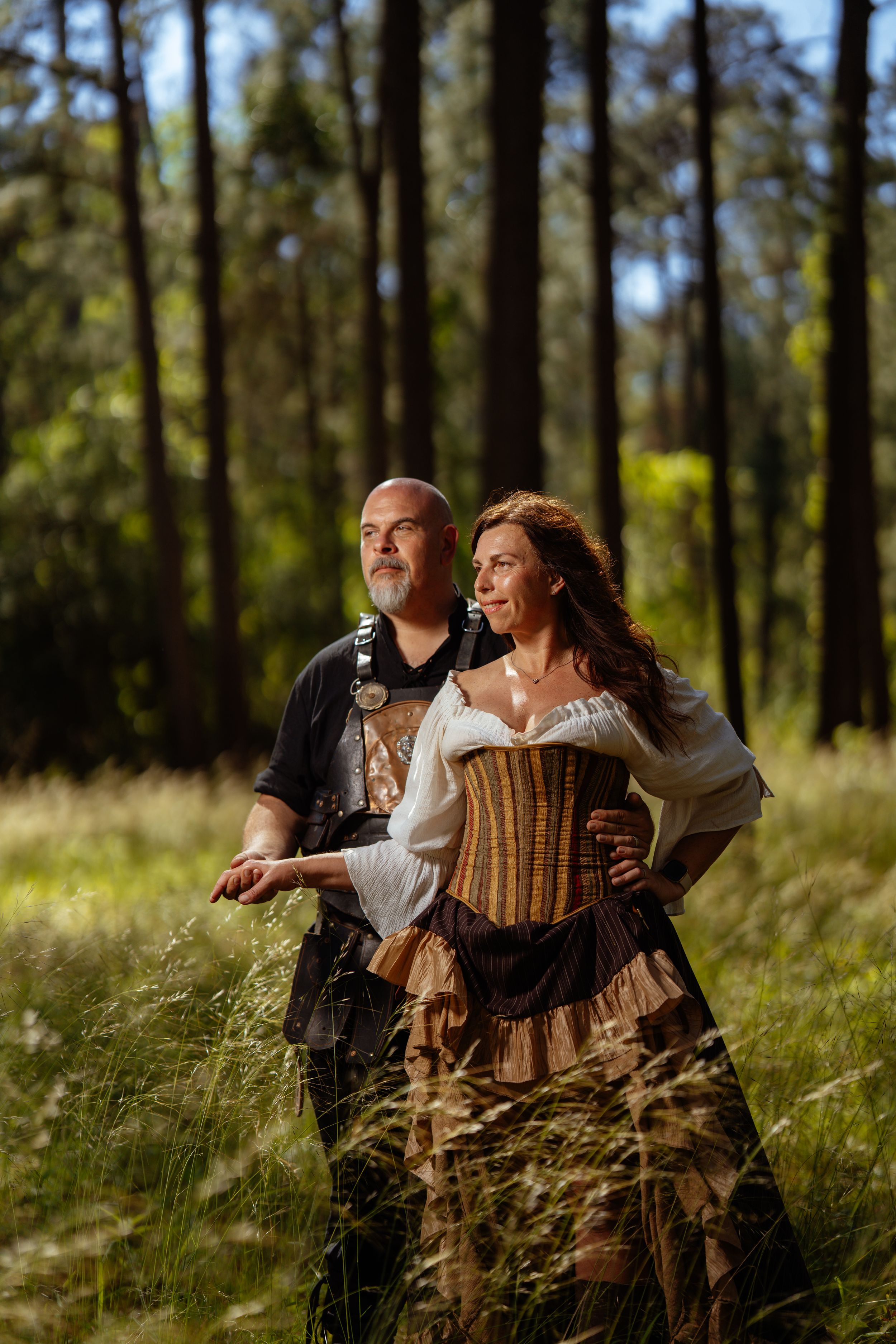 A couple in medieval or renaissance-inspired costumes poses together in a sun-dappled forest clearing.