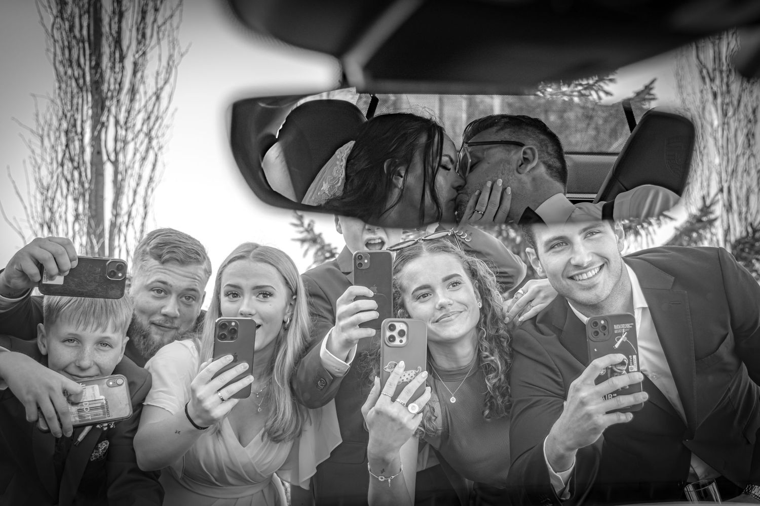 Black and white fun photo of wedding guests taking selfies through a car mirror.