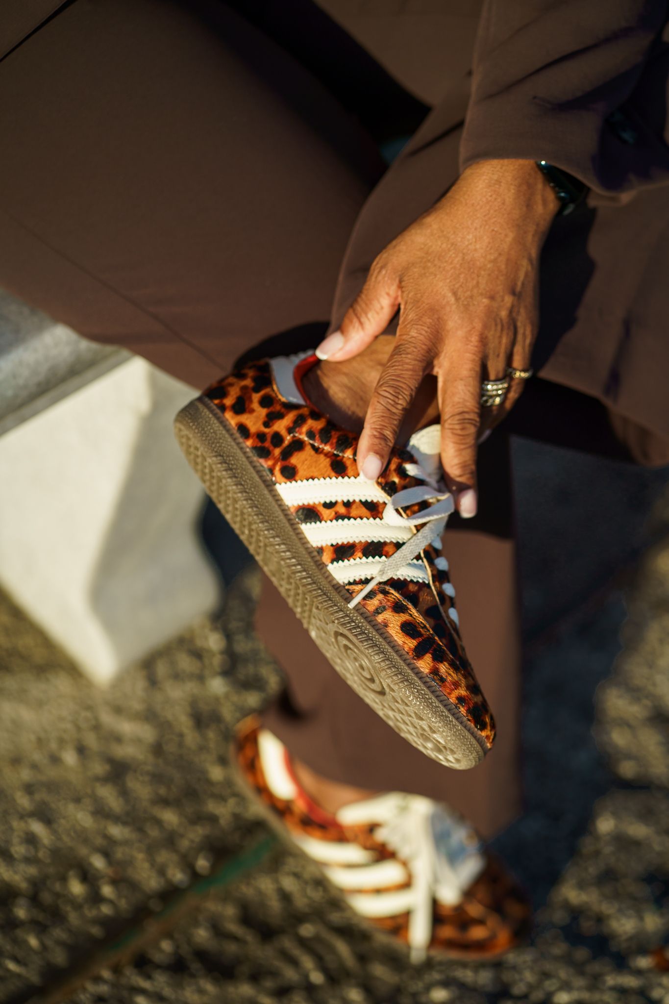 Person holding leopard print Adidas Samba sneakers with white stripes, wearing brown trousers outdoors.