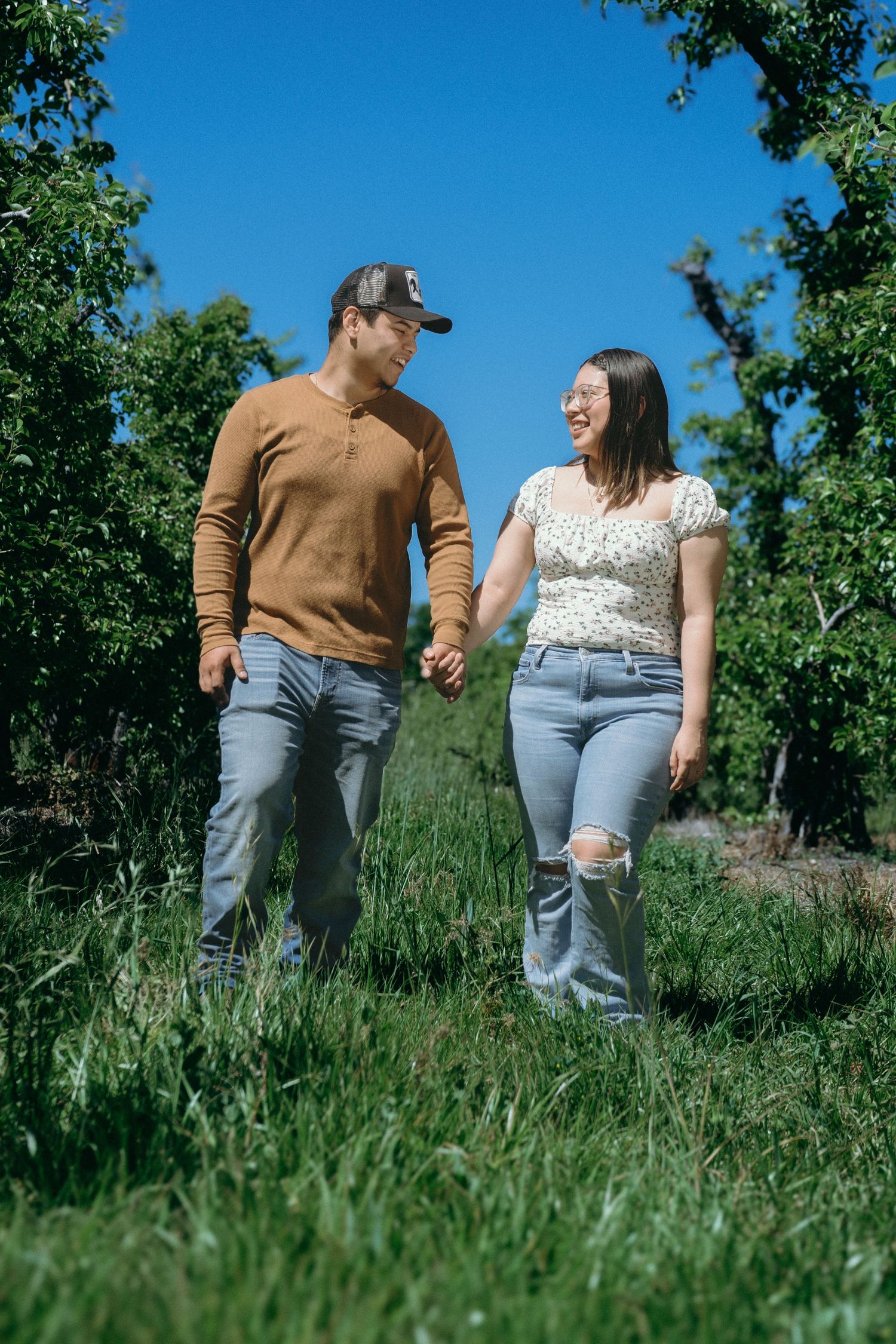 A happy couple holds hands while walking through a lush green orchard under a bright blue sky.