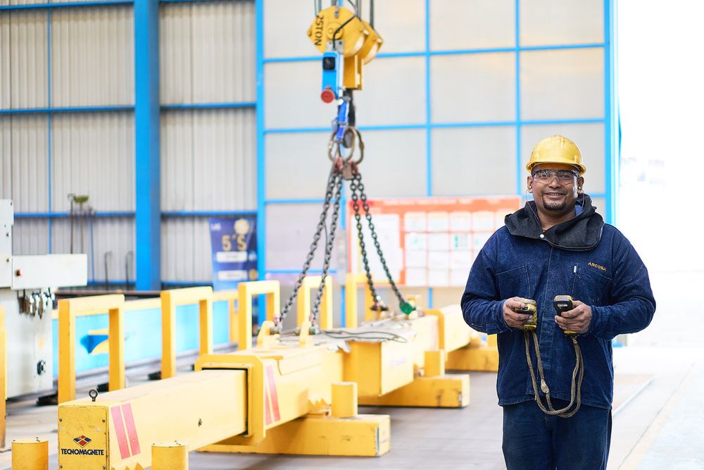Industrial worker in safety gear operates crane controls inside large manufacturing warehouse facility.