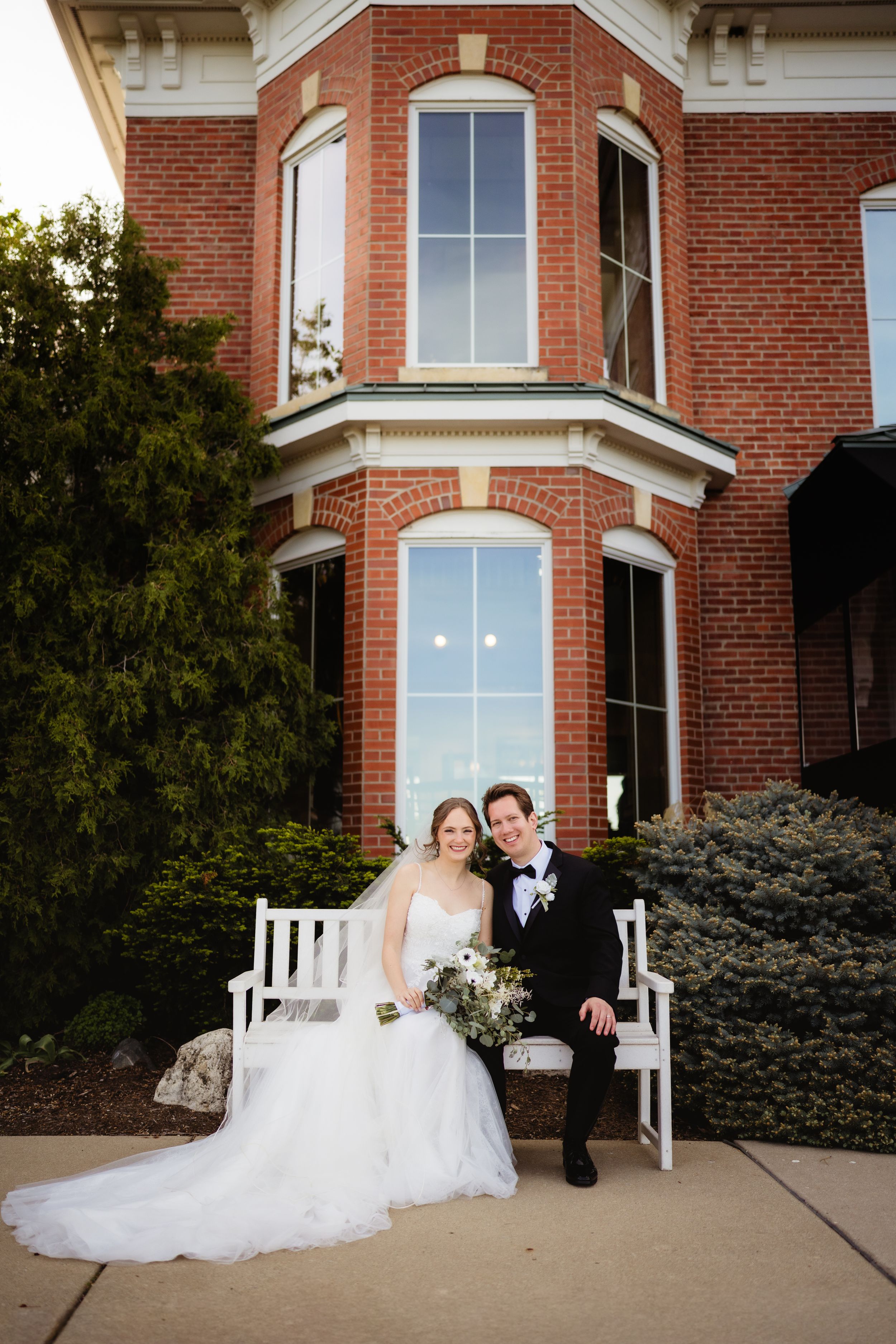 Bride and groom sit on white bench outside brick building, bride in white gown holding bouquet, groom in black tuxedo.