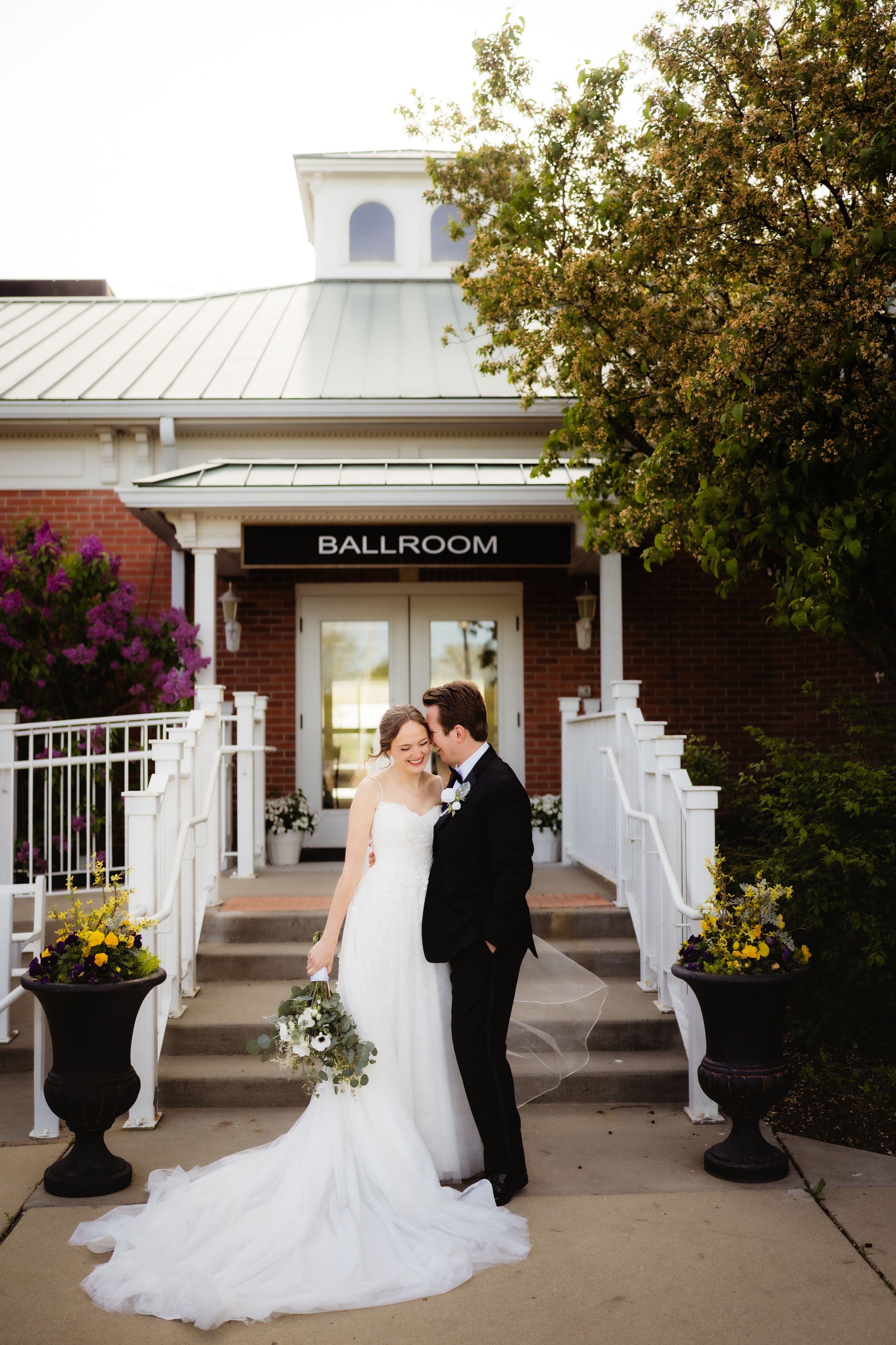 Bride and groom share a romantic moment outside a brick ballroom venue, surrounded by colorful flowers and lush greenery.