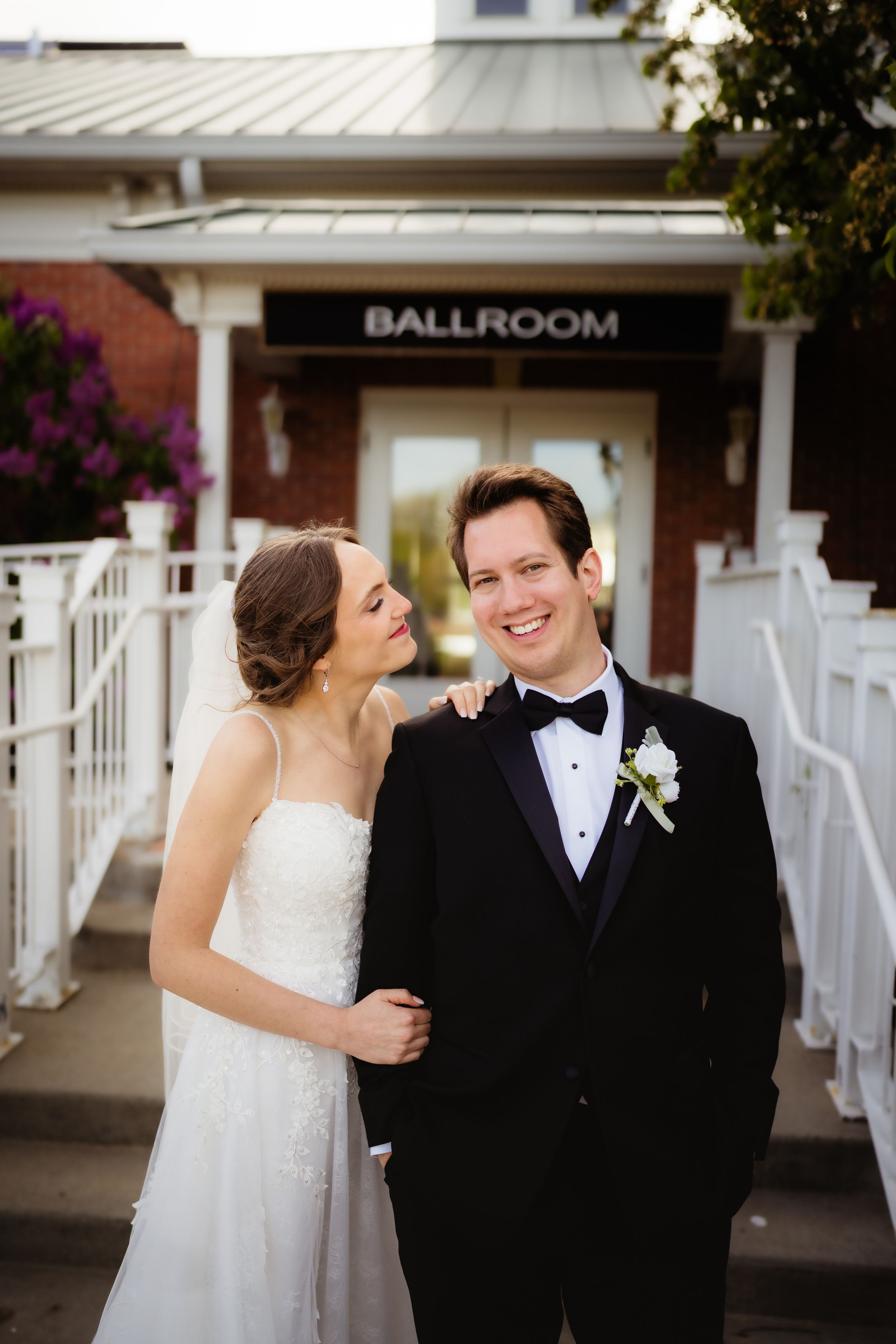 Bride in white gown and groom in black tuxedo smiling together outside a ballroom entrance on their wedding day.