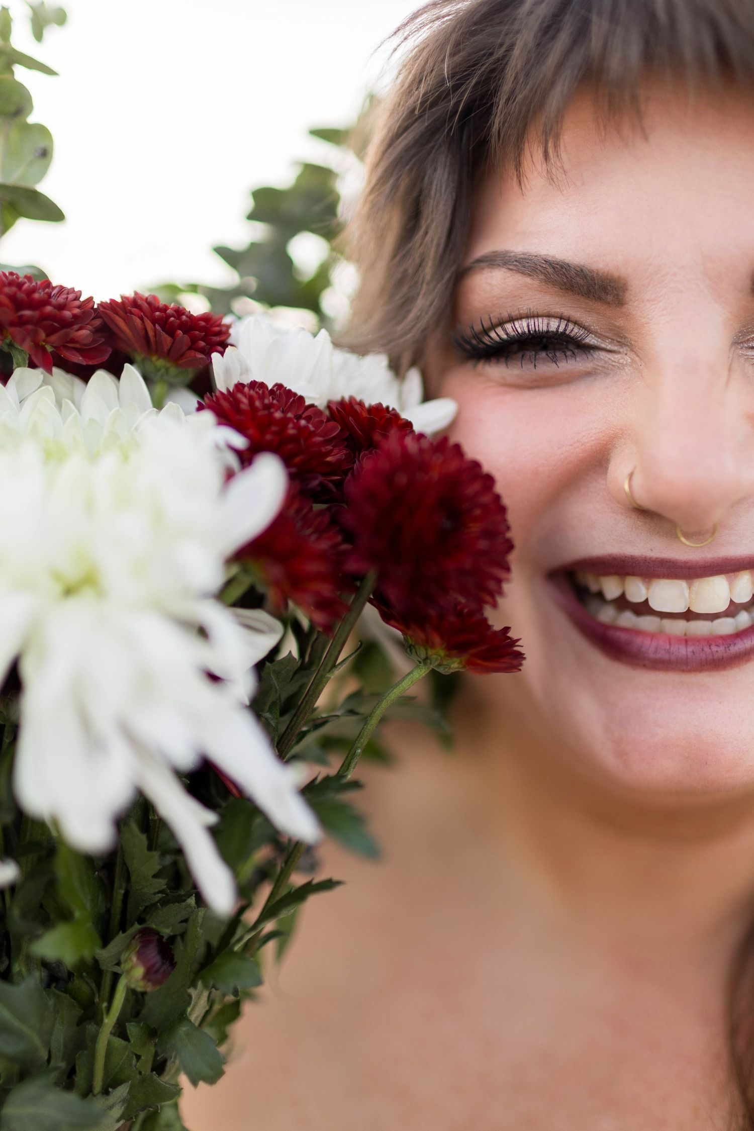 Smiling woman with short hair holding red and white chrysanthemum bouquet close to her face, wearing dark lipstick.