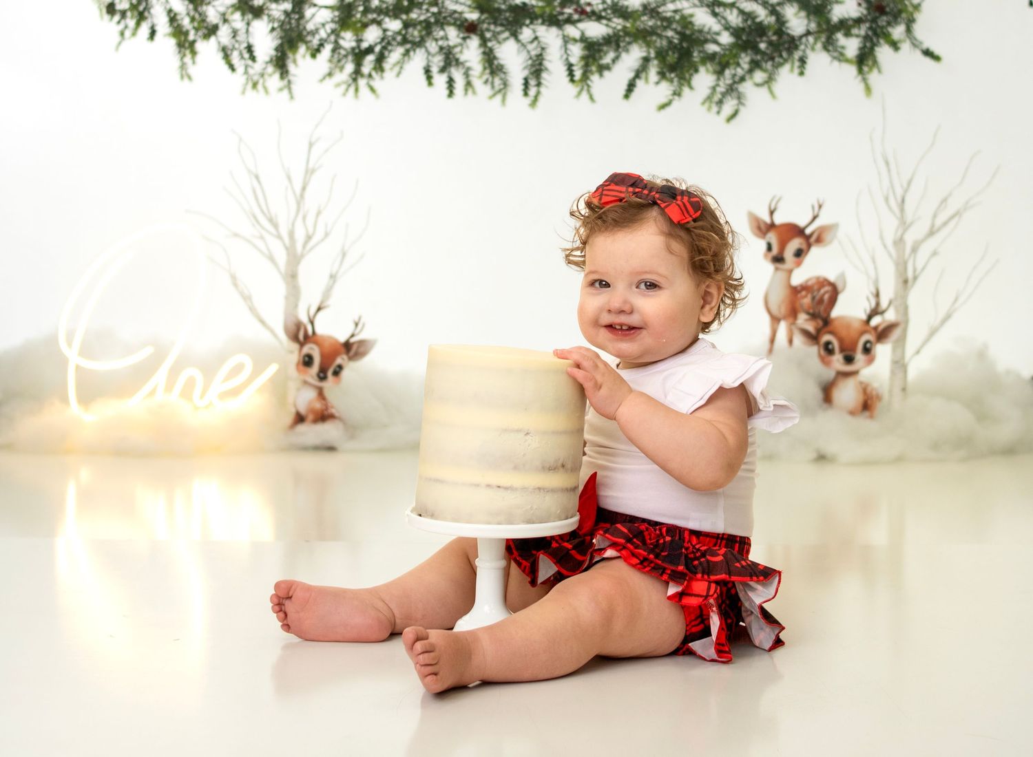 Smiling baby girl holds white cake in Christmas-themed cake smash session with reindeer and greenery backdrop.