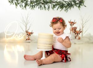 Baby girl smiles at her first birthday cake smash in a festive Christmas setting with reindeer decorations.