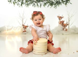 Baby girl smashing her first birthday cake in a festive Christmas-themed photo session with reindeer backdrop.