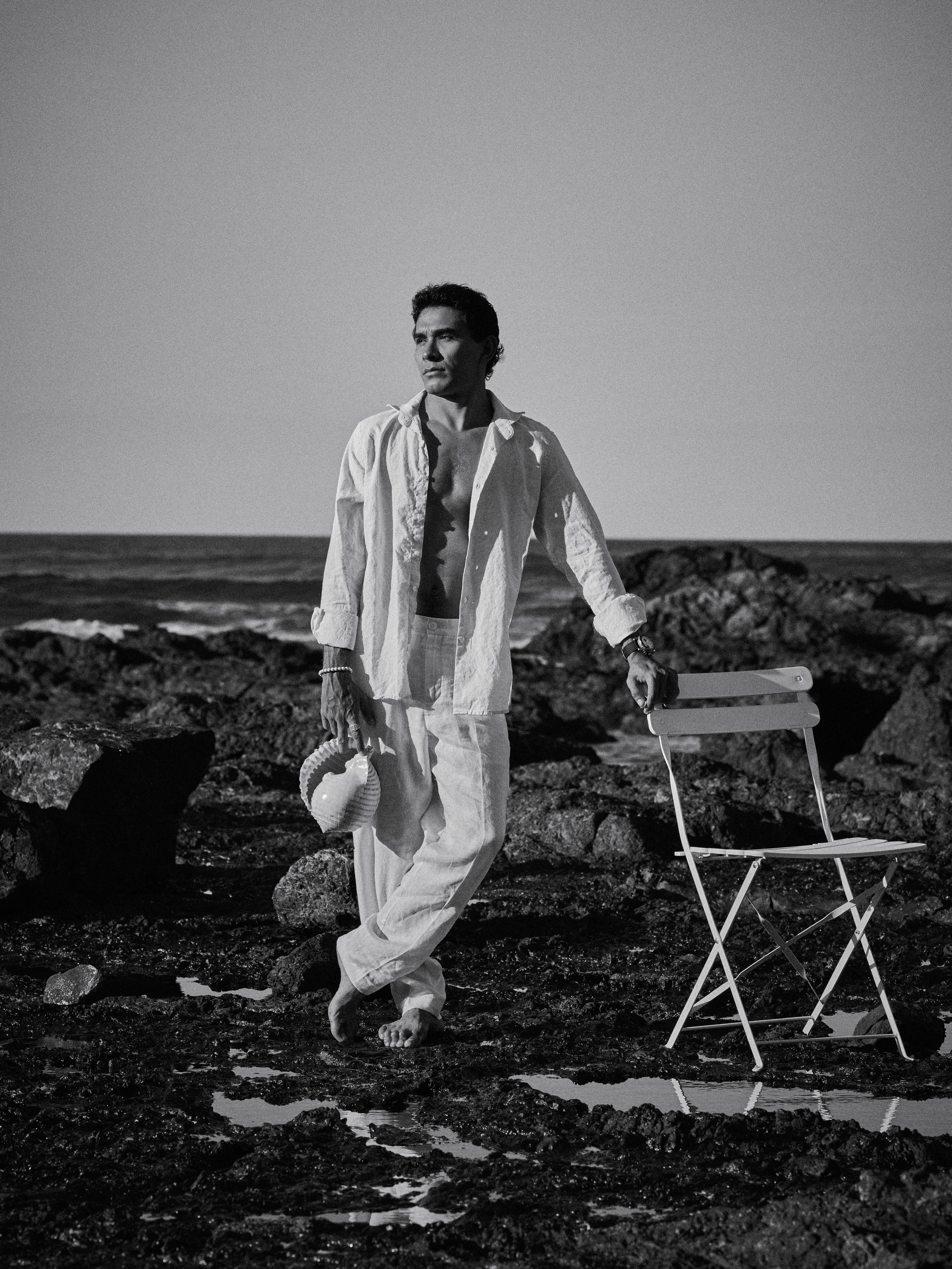 Man in white linen outfit stands by rocky shoreline next to folding chair, black and white photo.
