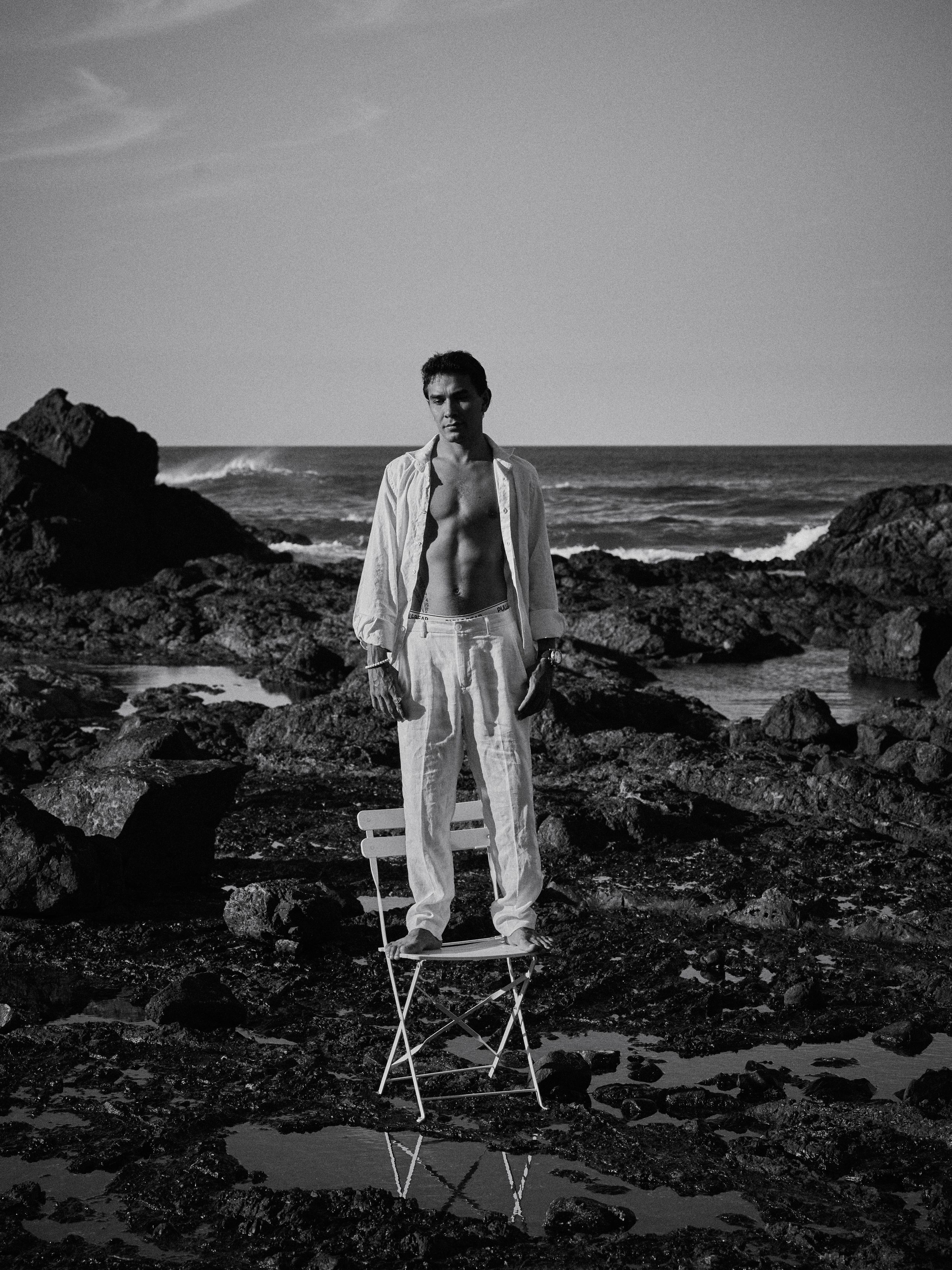 Black and white photo of a man in white linen outfit standing on a chair by rocky ocean shore.