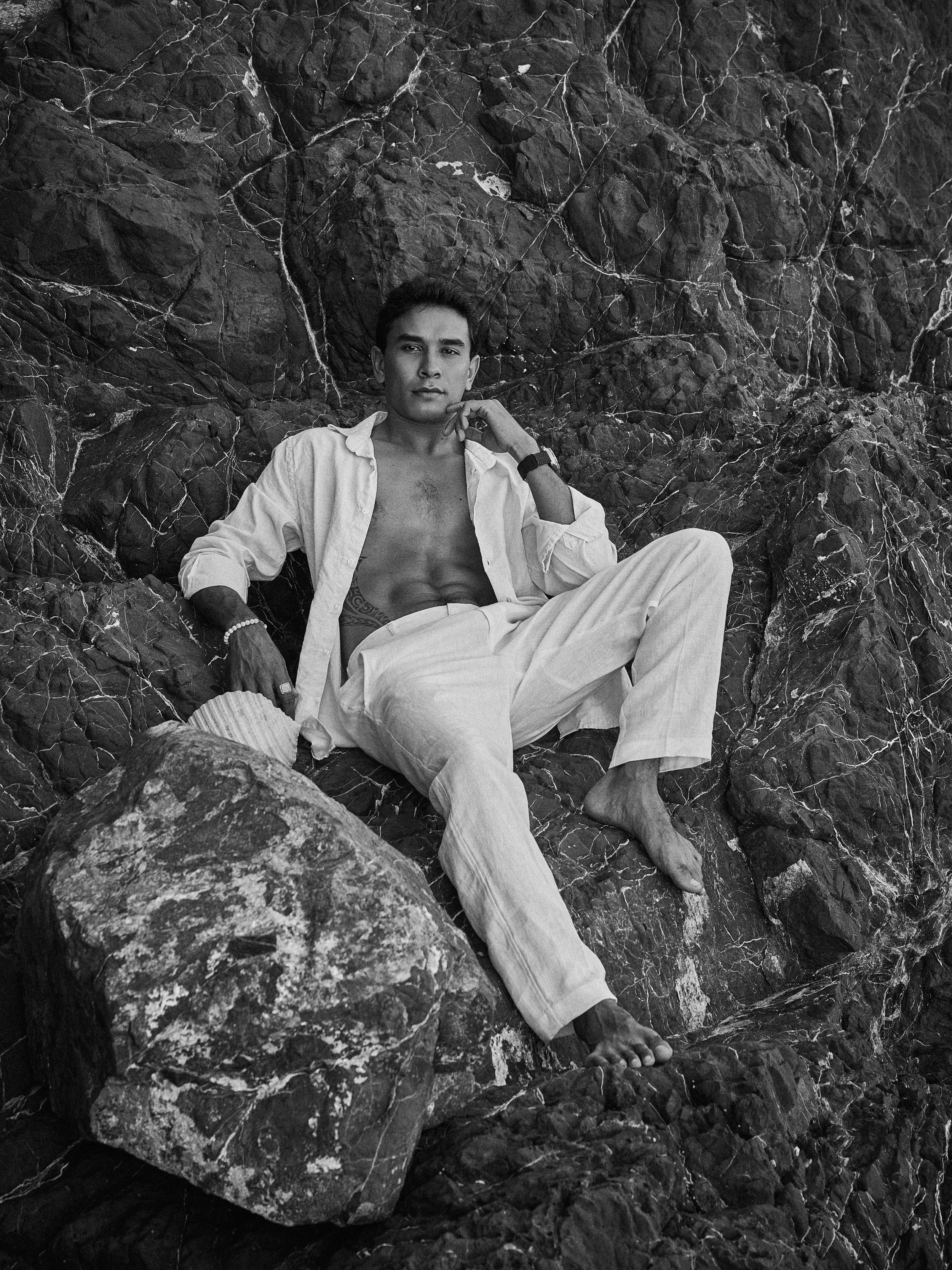 Black and white photo of a man in white linen outfit reclining on rocky coastal boulders.