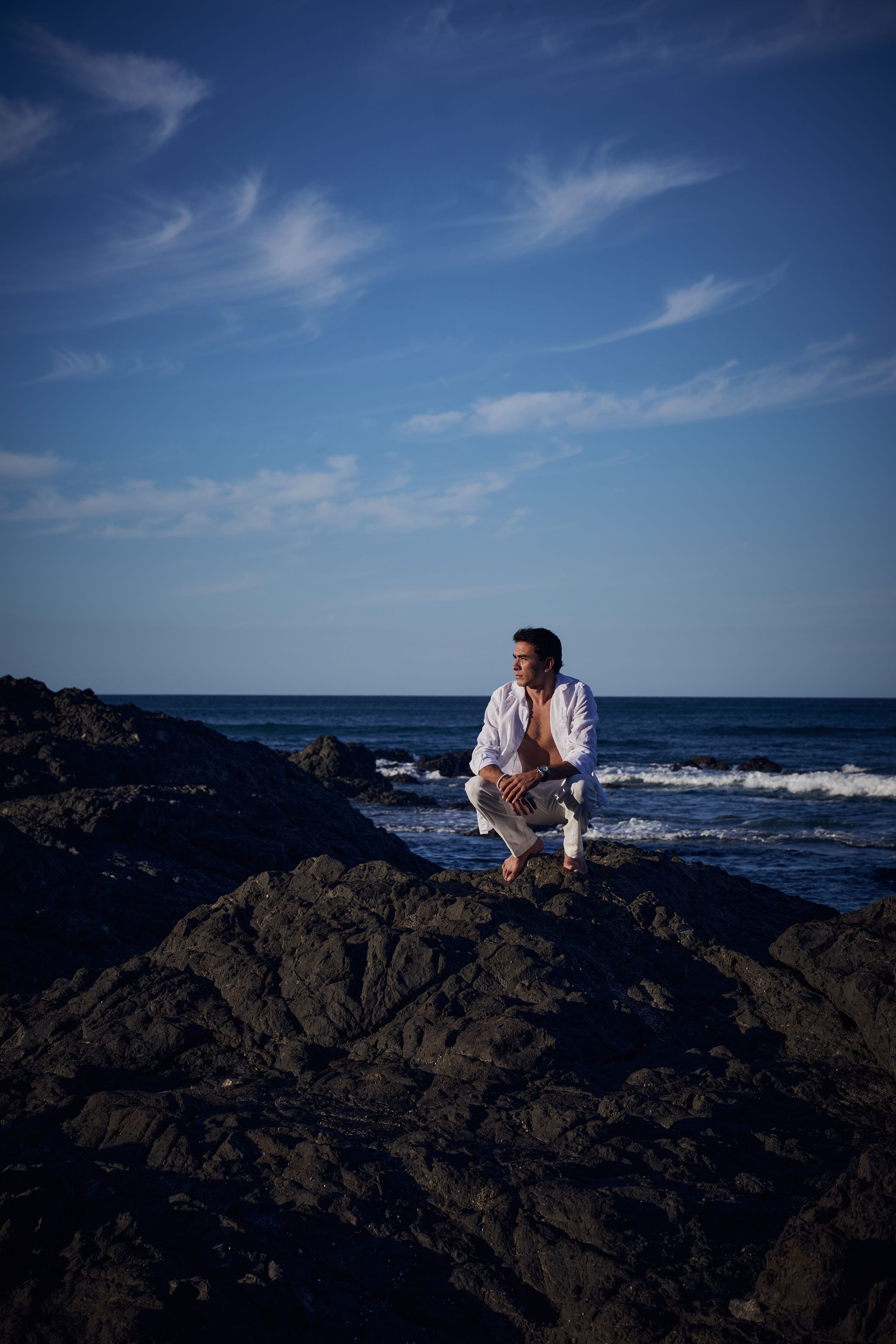 Man in white shirt crouching on dark rocks by the ocean under a blue sky with wispy clouds.