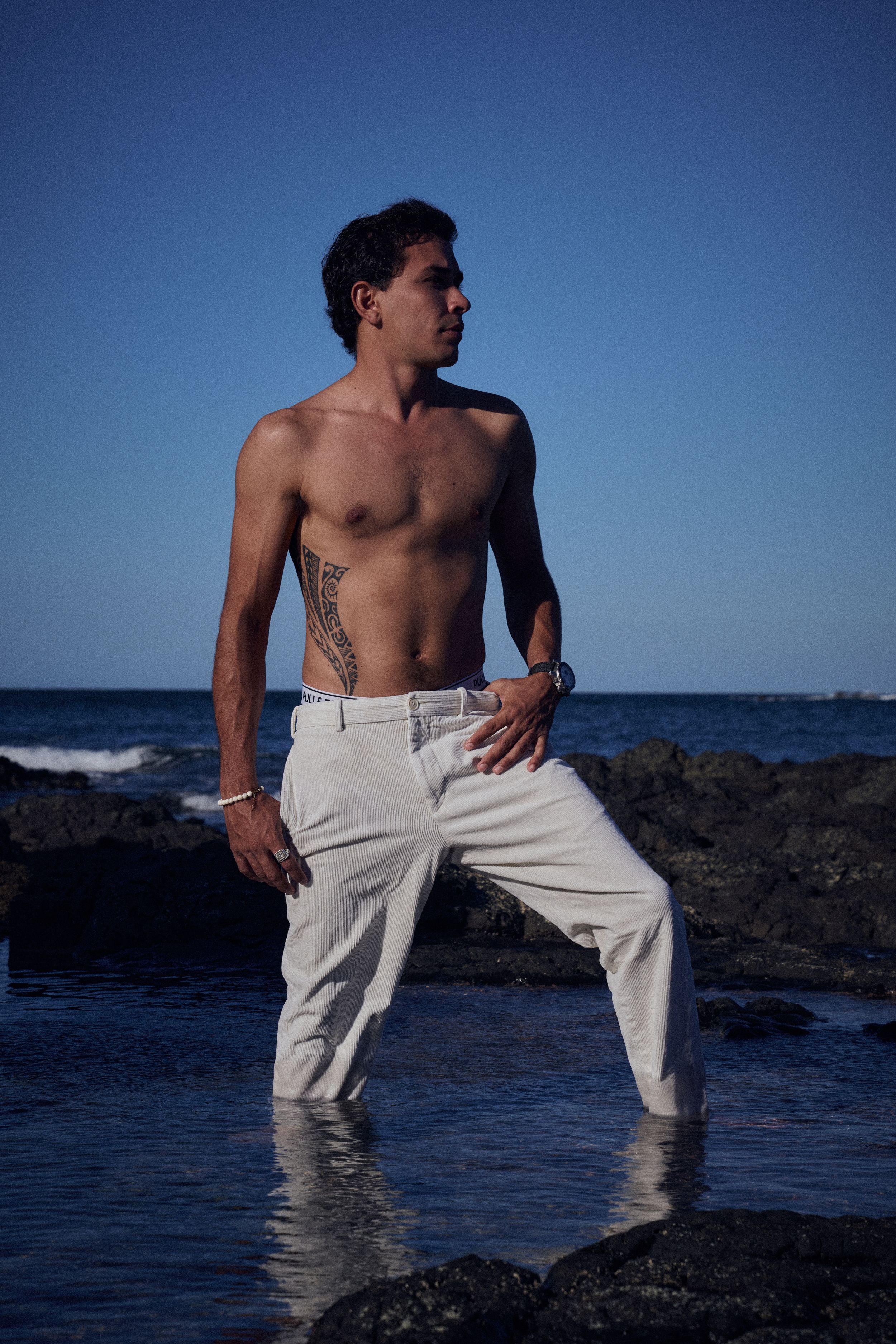 Shirtless man with tattoo in white pants standing in ocean water near rocks under clear blue sky.
