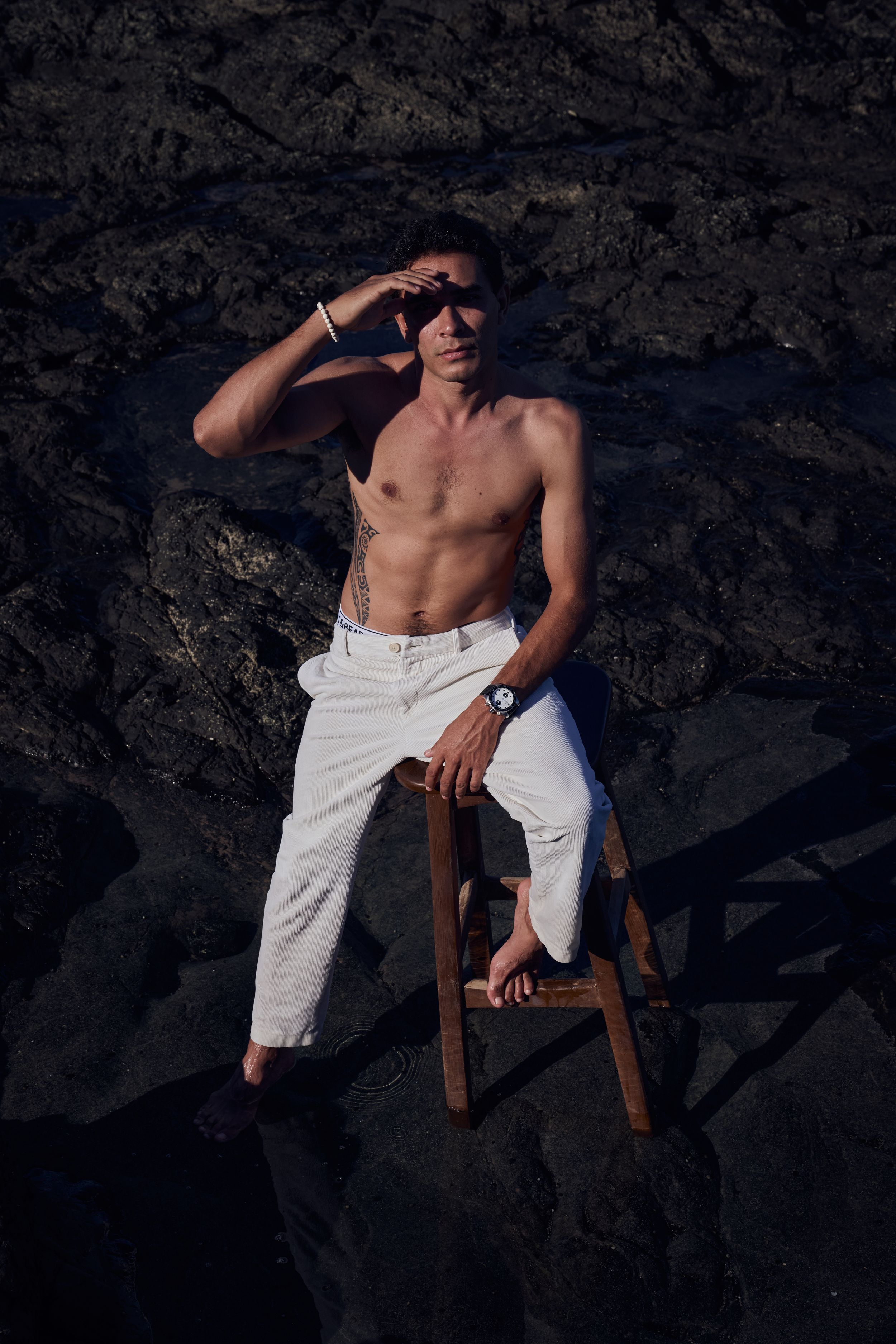 Shirtless man in white pants sitting on wooden stool against dark rocky background, shielding eyes from sun.