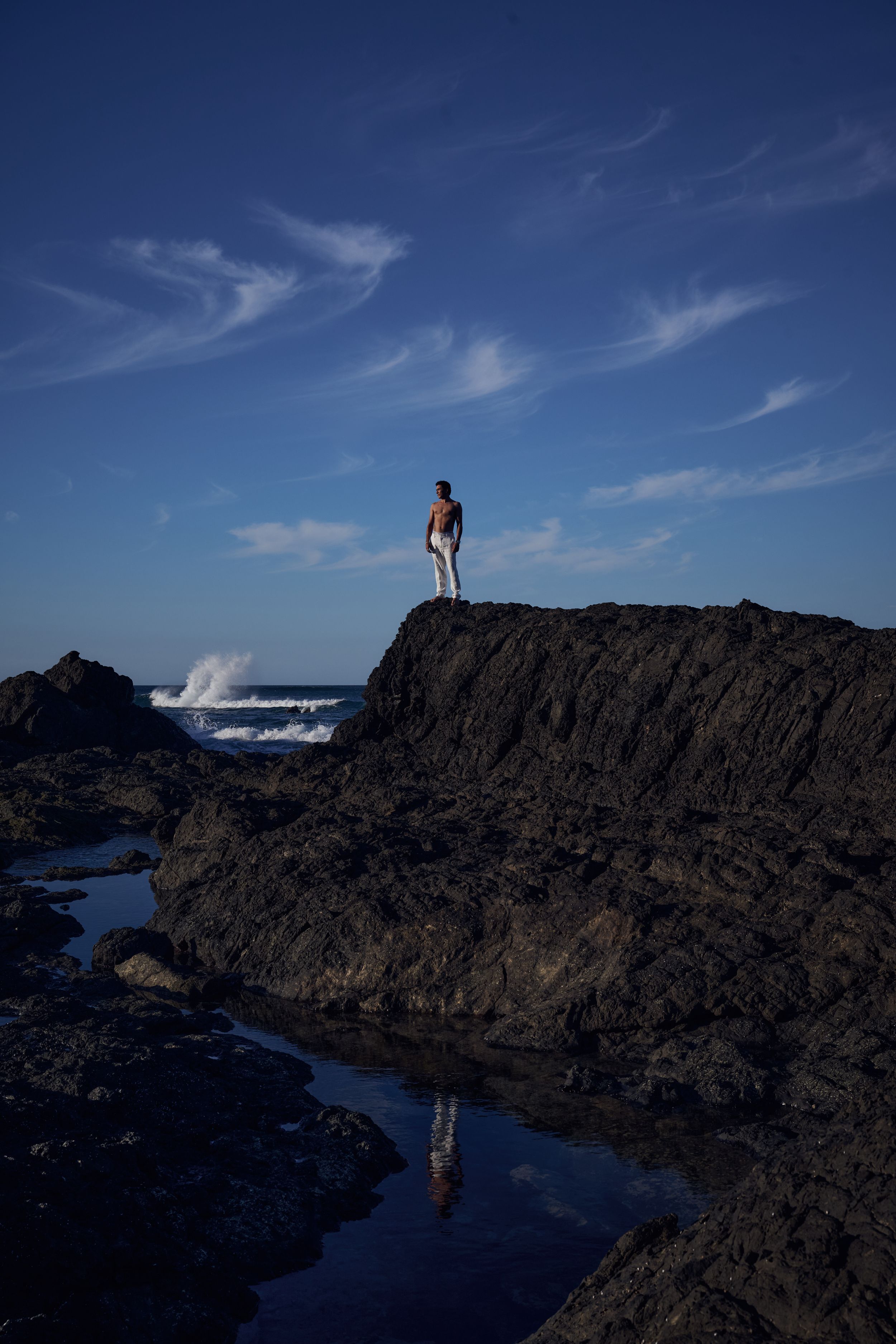 A shirtless man stands atop dark coastal rocks overlooking crashing ocean waves under a deep blue sky.